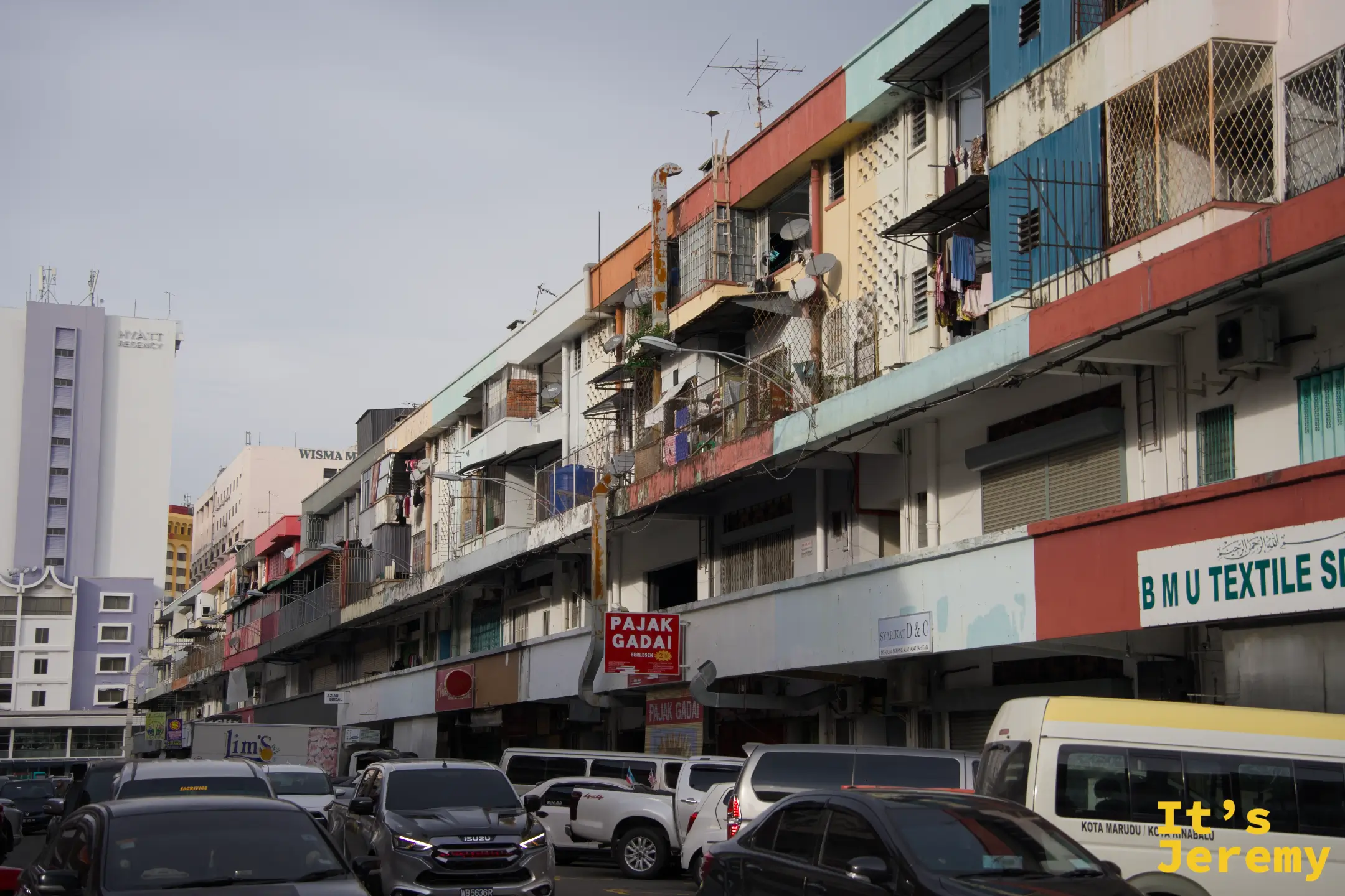 亞庇老街區街景，舊式騎樓商店街道，車輛穿梭，招牌林立，PAJAK GADAI 當鋪標誌可見