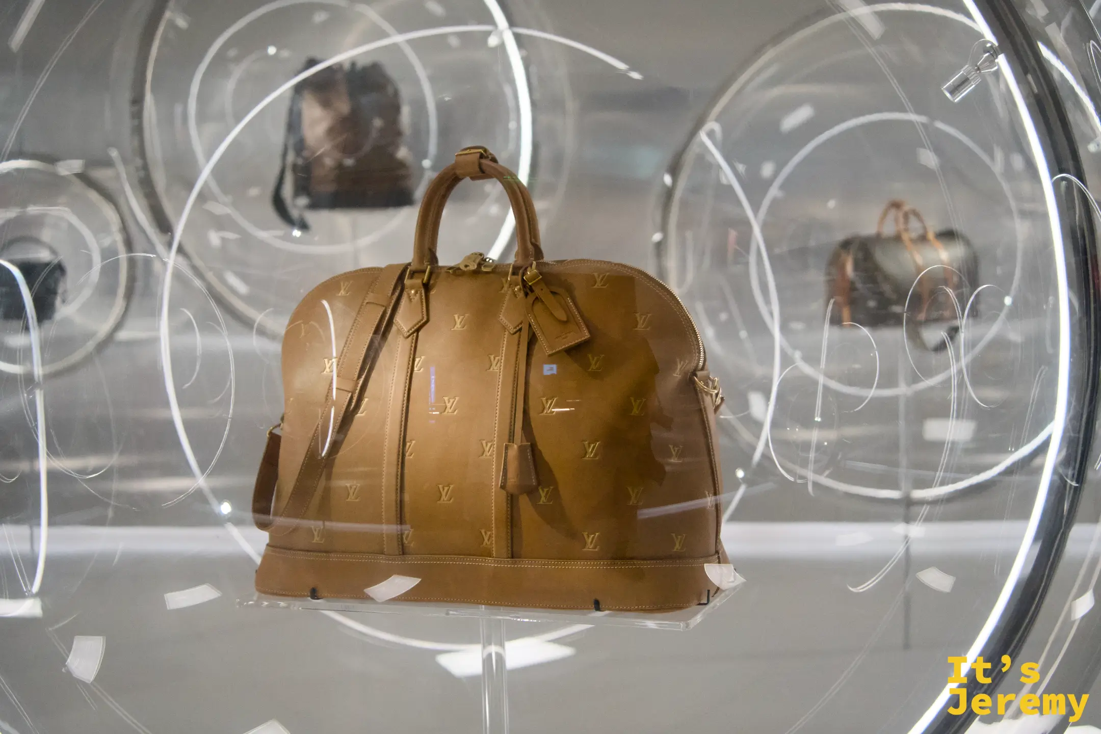 圓形壓克力展示泡泡，中央是棕色 monogram 皮革 Keepall 旅行包，聚光燈透過球面折射出柔和光暈