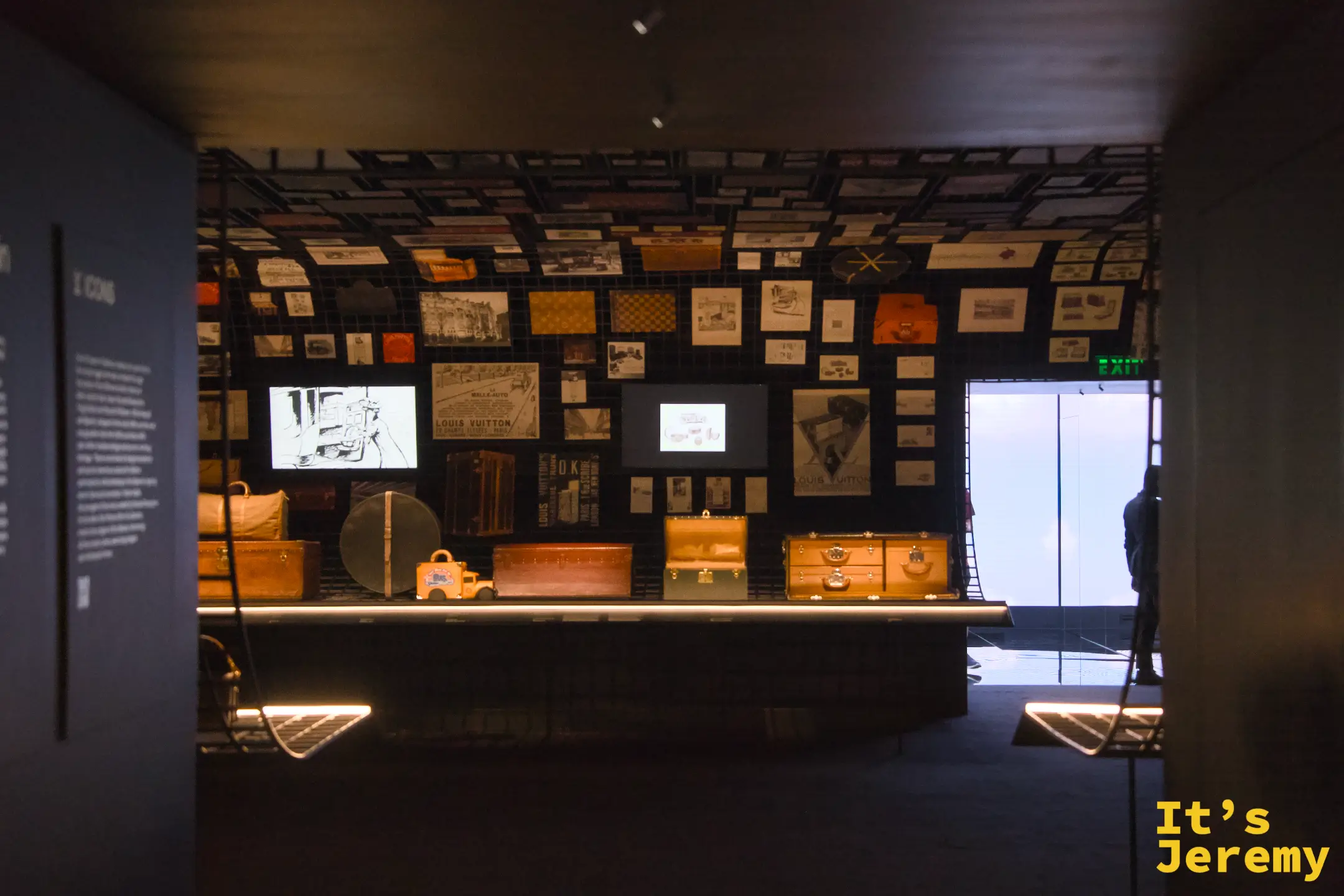 Origins 歷史檔案室全景，展台上排列多款不同年代行李箱，背景大螢幕與滿牆老廣告形成強烈縱深