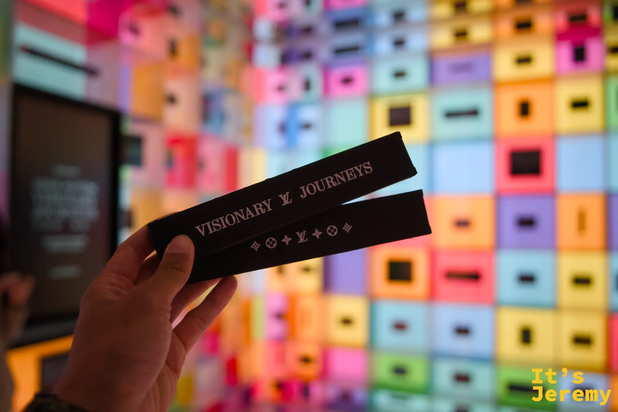 扭蛋機送出的鉛筆組紀念品，黑底燙印「VISIONARY LV JOURNEYS」字樣，背景是多彩格子燈箱牆