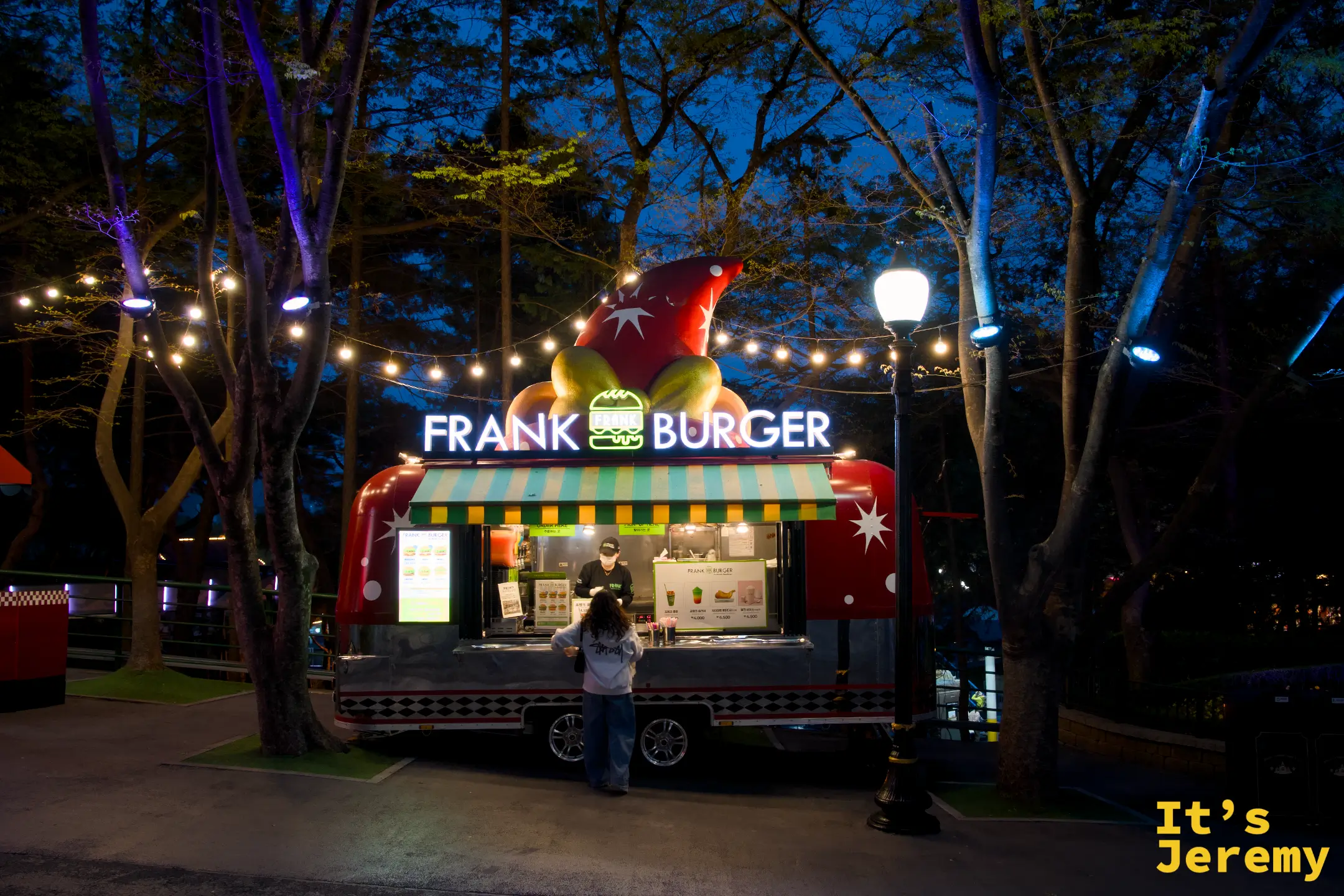 Frank Burger 餐車，紅色木屋造型車身、車頂是漢堡蔬菜造型雕塑，白底霓虹「FRANK BURGER」招牌，前方掛著一串黃色小燈泡，一位戴帽子的人站在窗口前