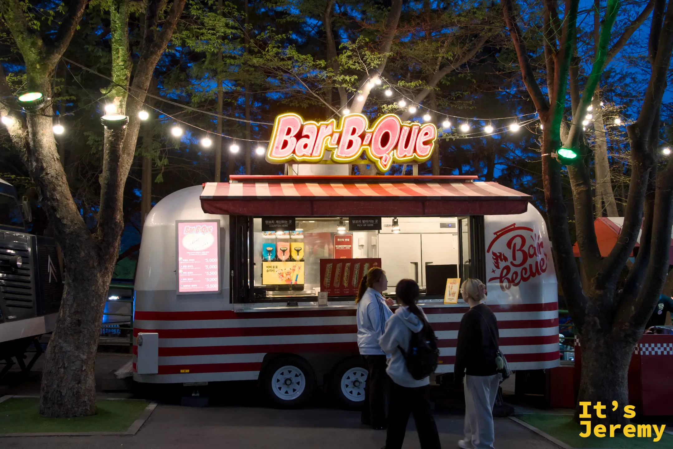 Bar-B-Que 餐車，銀白色復古車身、白底紅條紋帆布頂篷，車頂掛著黃紅色霓虹「Bar-B-Que」招牌，三位女生站在窗口前點餐，車頂兩側拉著一串小串燈
