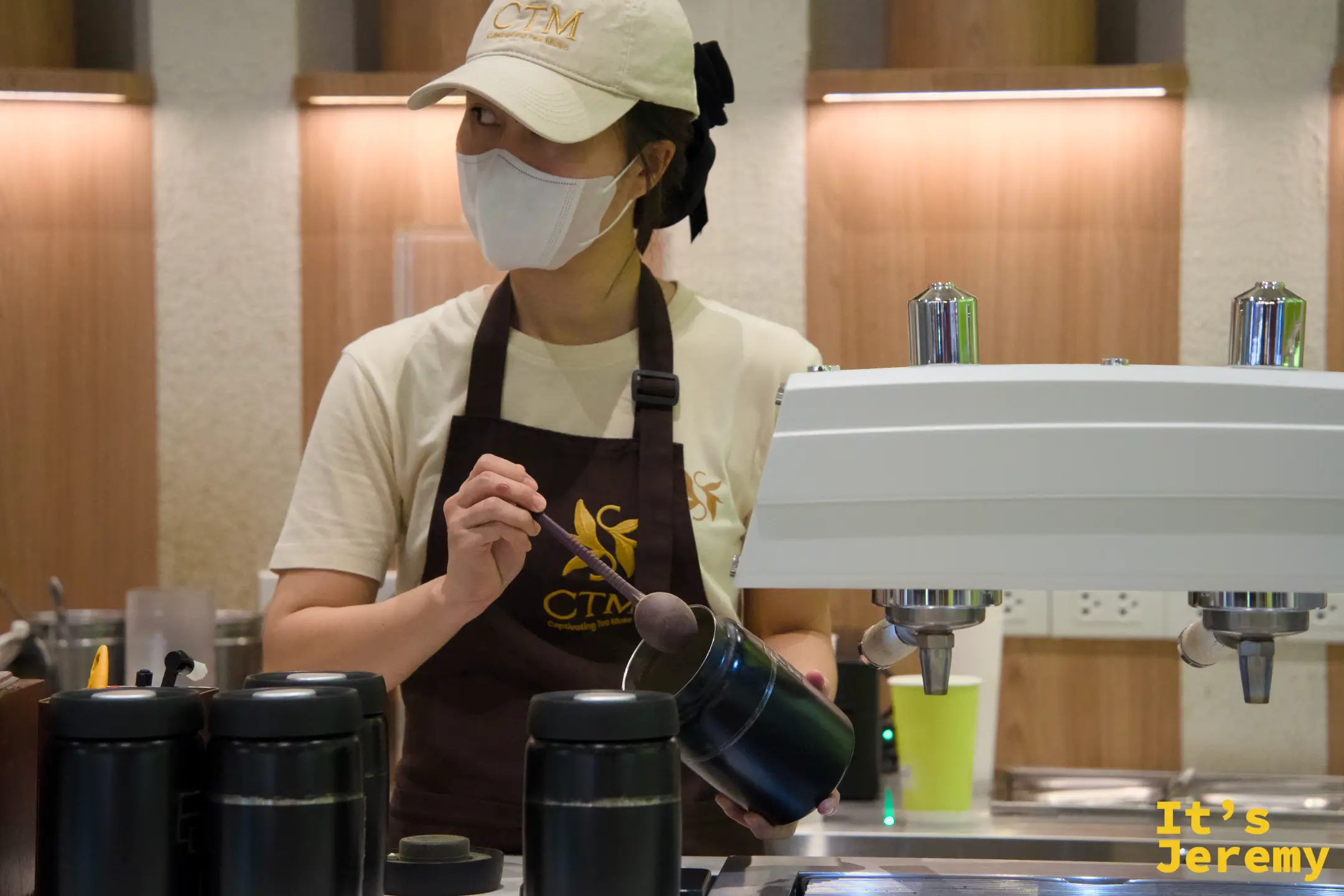 女性店員攪拌拿鐵，深色圍裙上繡有 CTM 花葉 logo
