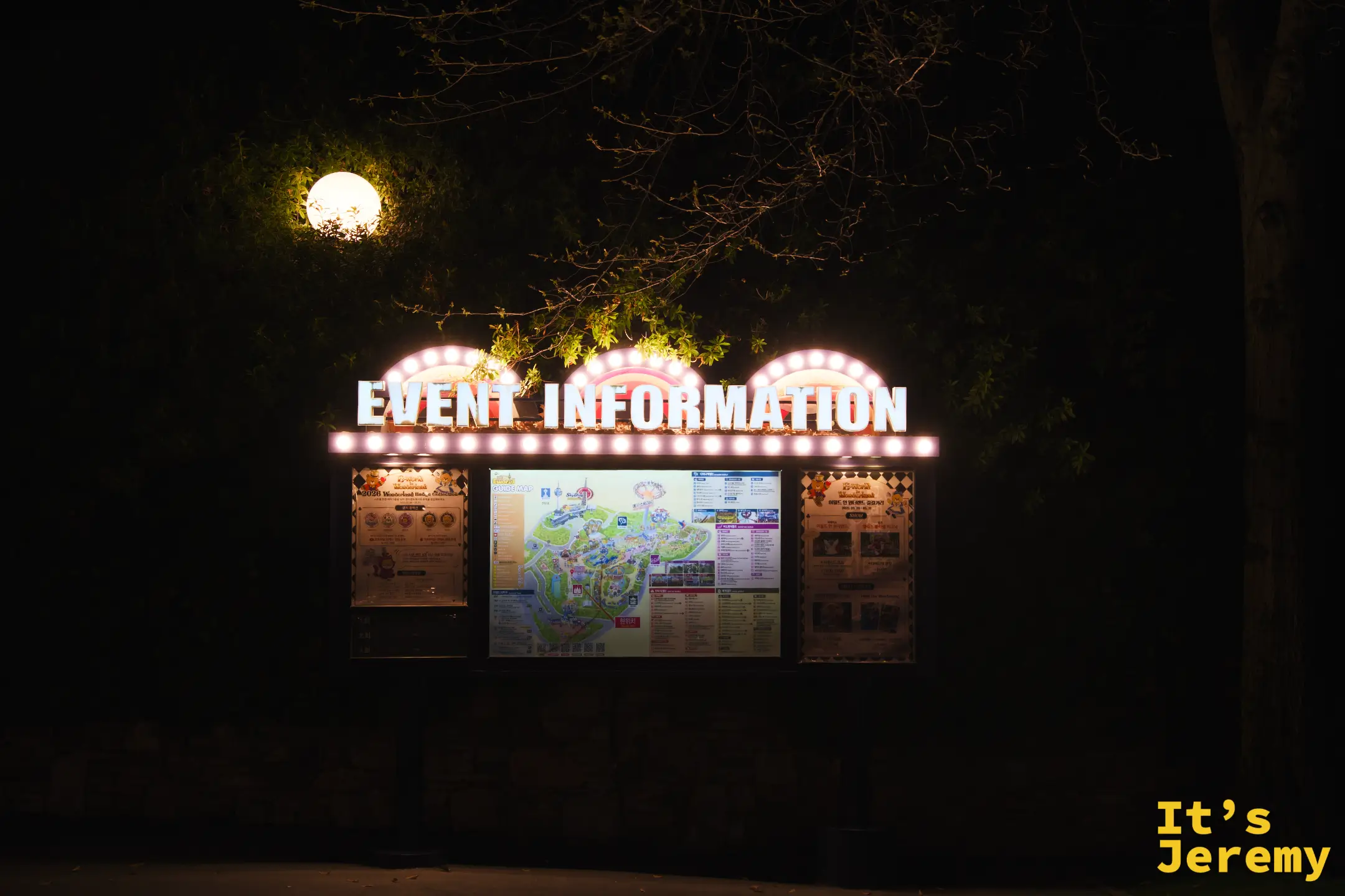 Event Information 公告牌：頂部三個拱形邊框鑲滿黃色小燈泡，下方三塊看板印著園區地圖與當日活動表，背景是深色樹影與一盞發光的球形路燈