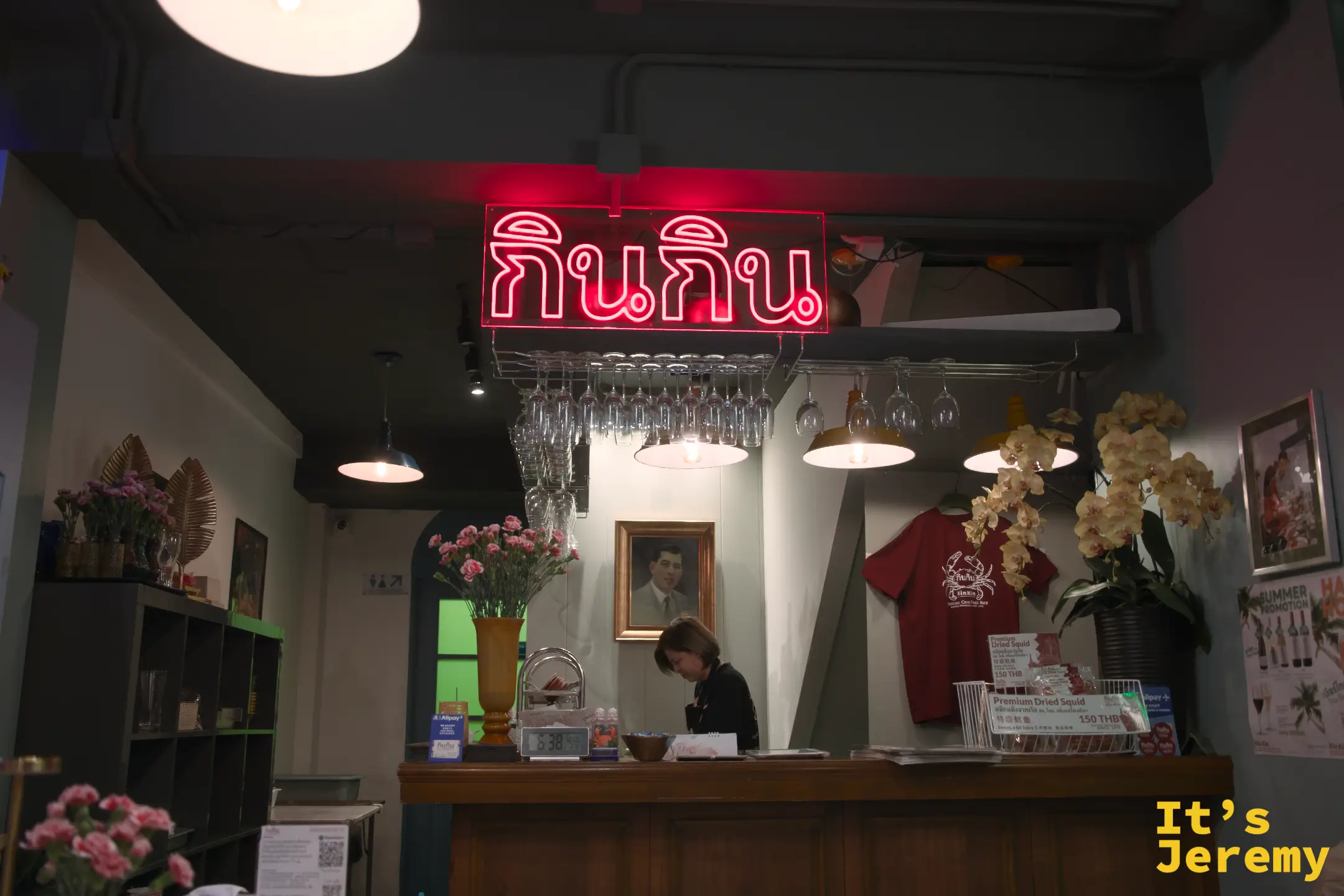 吧台上方的紅色霓虹「กินกิน」字樣，後方掛著一幅老照片，收銀台旁展示著自家品牌周邊