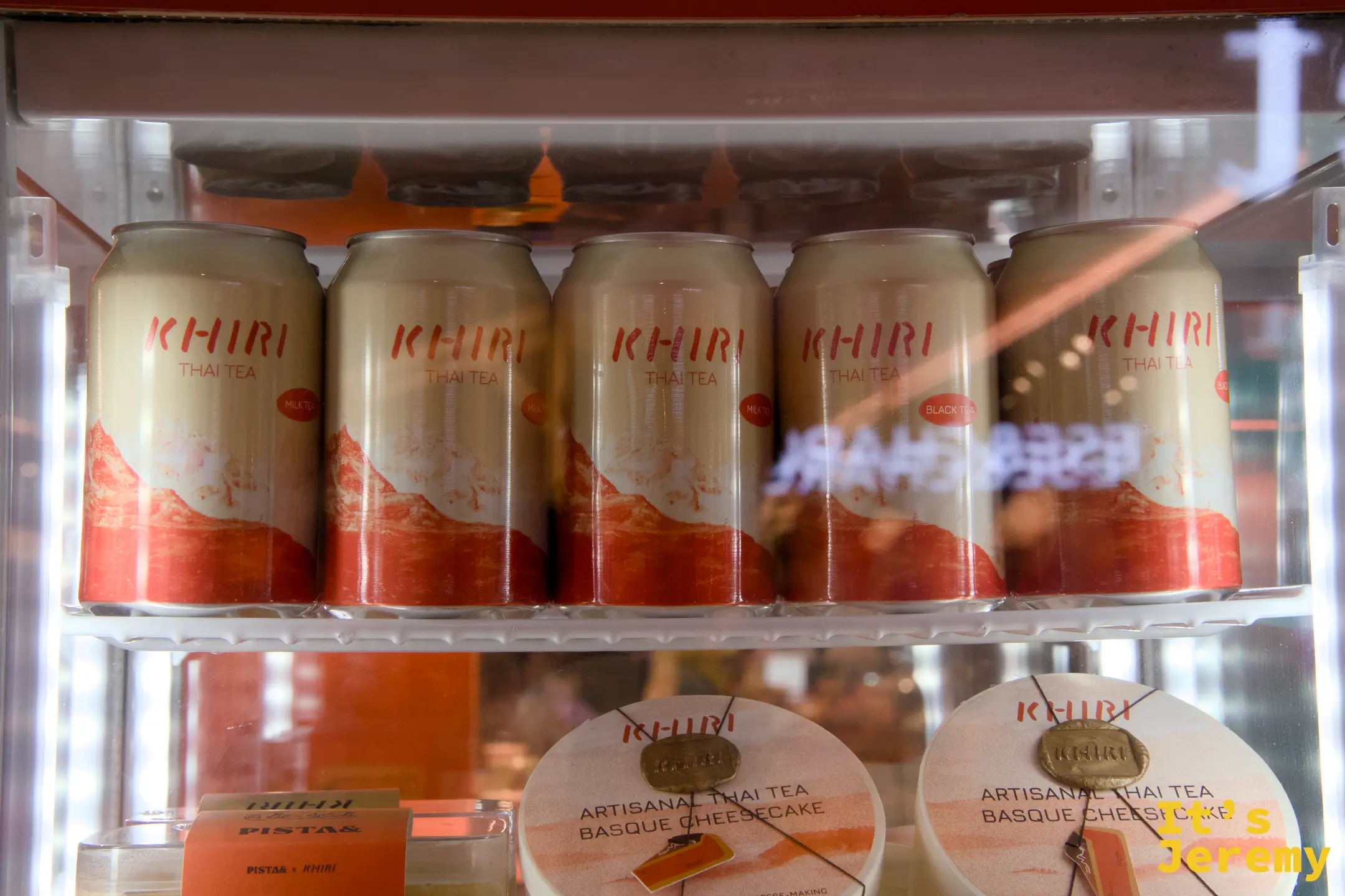 展示冰箱近景:橘色 KHIRI 罐裝(Milk Tea 與 Black Tea 款)與 Artisanal Thai Tea Basque Cheesecake