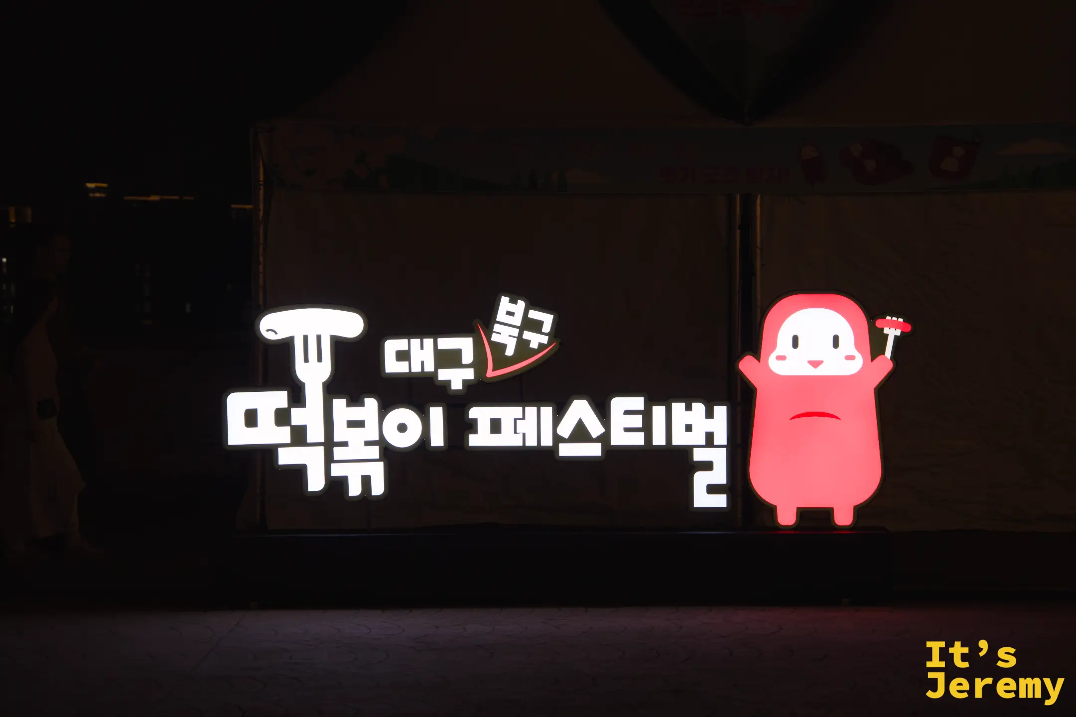 大邱북구떡볶이페스티벌（大邱北區辣炒年糕節）的發光招牌，黑色帳篷前的白底字樣與右側橘色卡通吉祥物（手拿叉子與年糕）發光跳出畫面