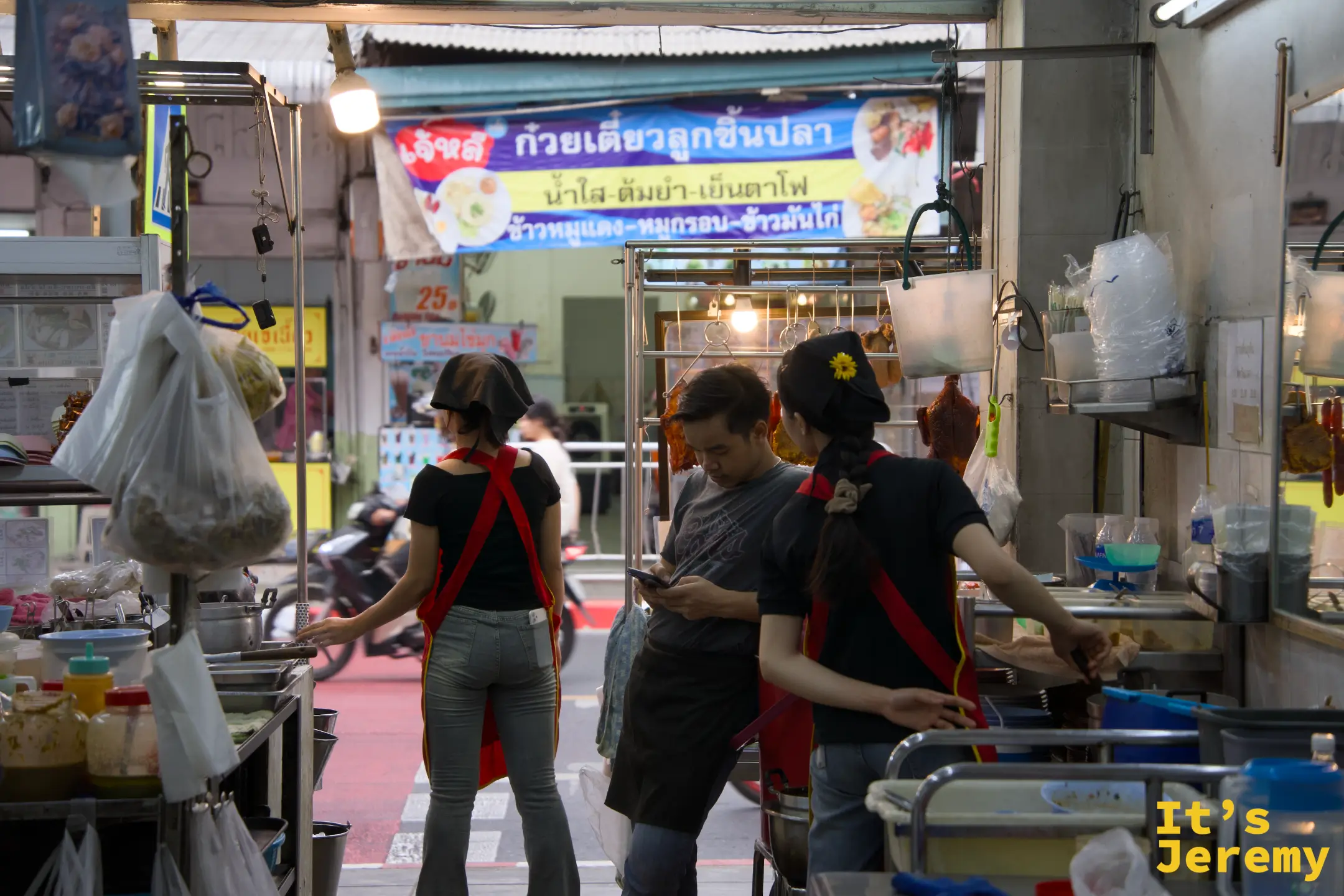 店內景象:穿黑衣紅圍裙的服務員和客人在點餐,上方是泰文招牌「เจ้าหลงก๊วยเตี๋ยวลูกชิ้นปลา」