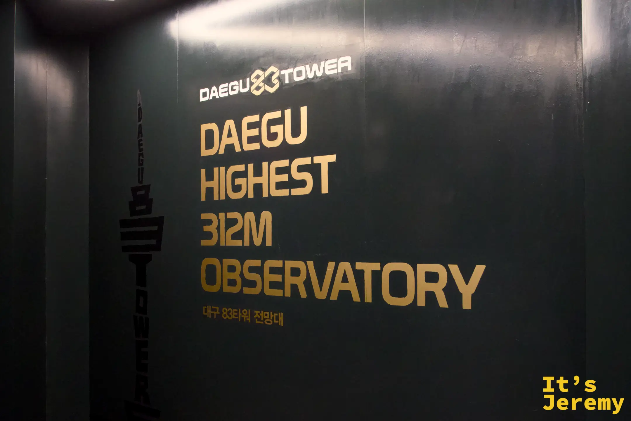 83 塔觀景台入口的深綠色牆面，正中央印著金色英文「DAEGU HIGHEST 312M OBSERVATORY」與底下小字韓文「대구 83타워 전망대」，牆面左側有 Daegu Tower 的塔身剪影標誌