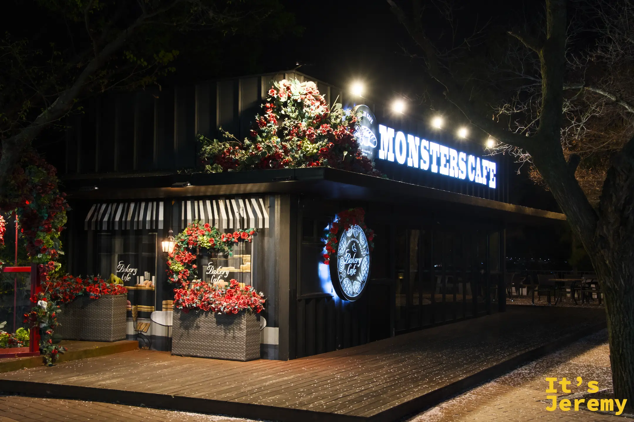 Monsters Cafe 外觀，黑色貨櫃屋造型咖啡店，藍色霓虹「MONSTERS CAFE」招牌，門口被大量紅花綠葉裝飾包圍，圓形「Bakery Cafe」標誌掛在門邊