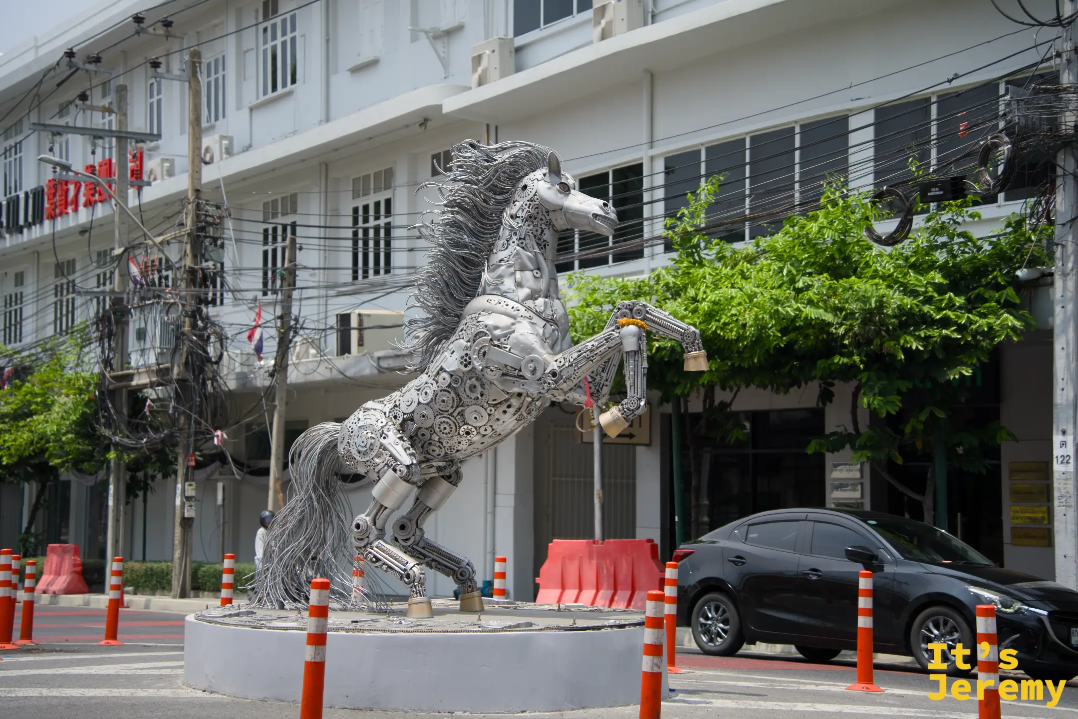 路口擺放的大型廢鐵件拼組金屬馬雕塑,揚蹄姿態,後方是白色建築