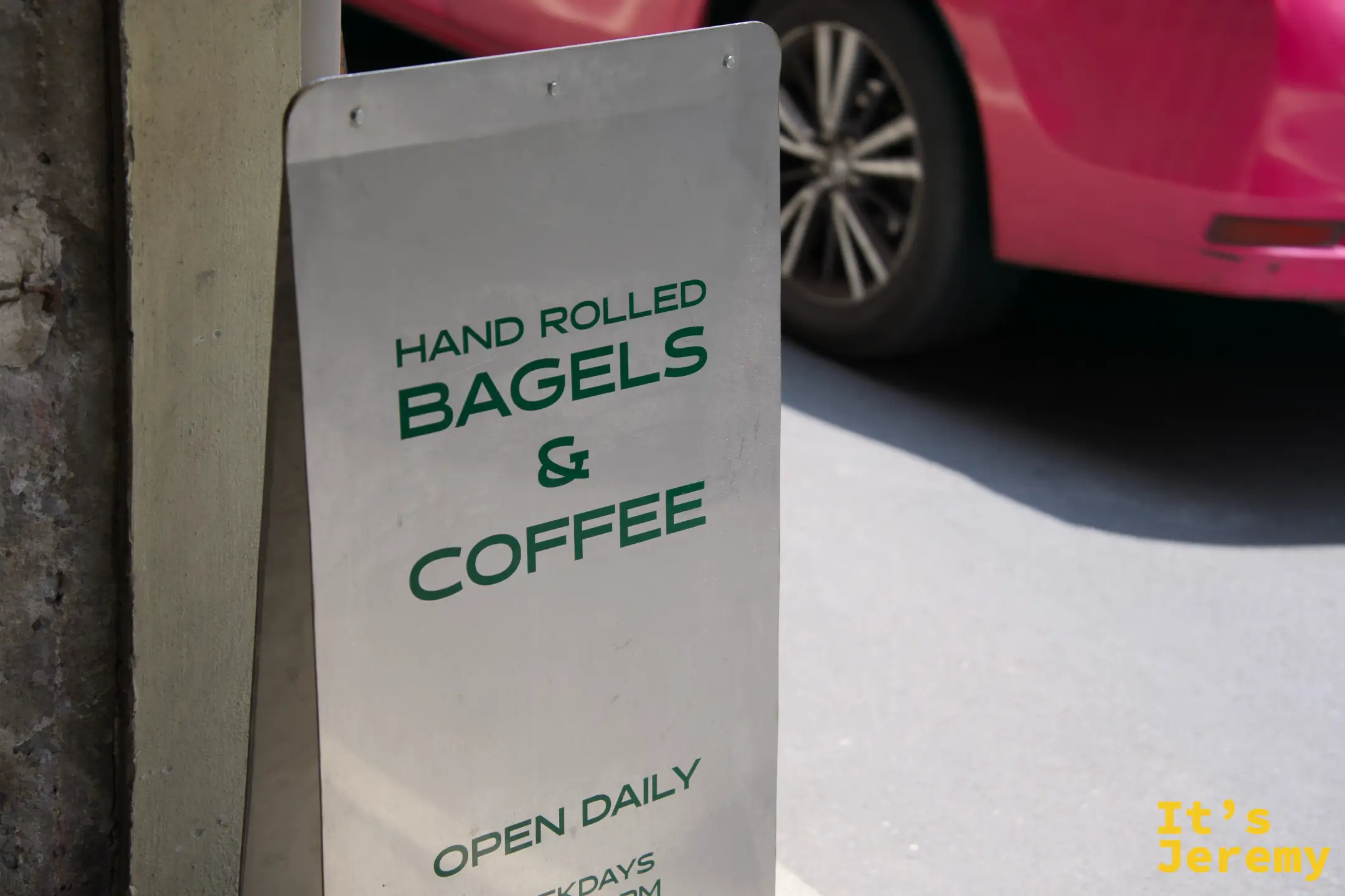 店門口的金屬立牌:「HAND ROLLED BAGELS & COFFEE, OPEN DAILY」,背景是路邊停放的紅色汽車