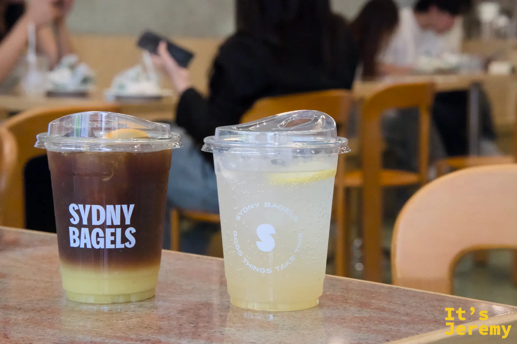 桌面上的兩杯飲料:「SYDNY BAGELS」字樣的黑咖啡與印有圓形 logo 的氣泡飲並排