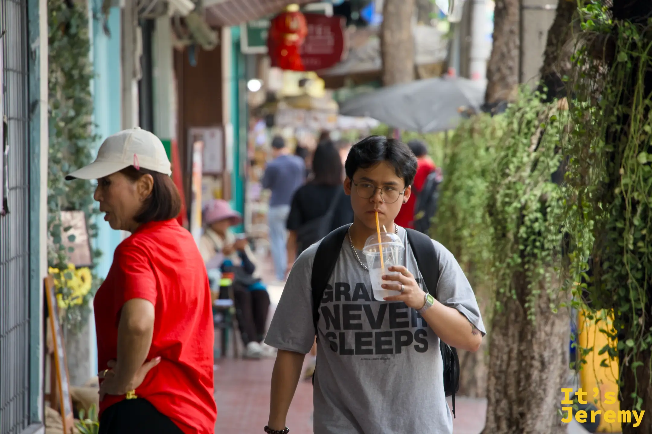 年輕男子邊走邊用吸管喝飲料,T 恤印著「Grains Never Sleeps」,背後是懸掛植物的廊道
