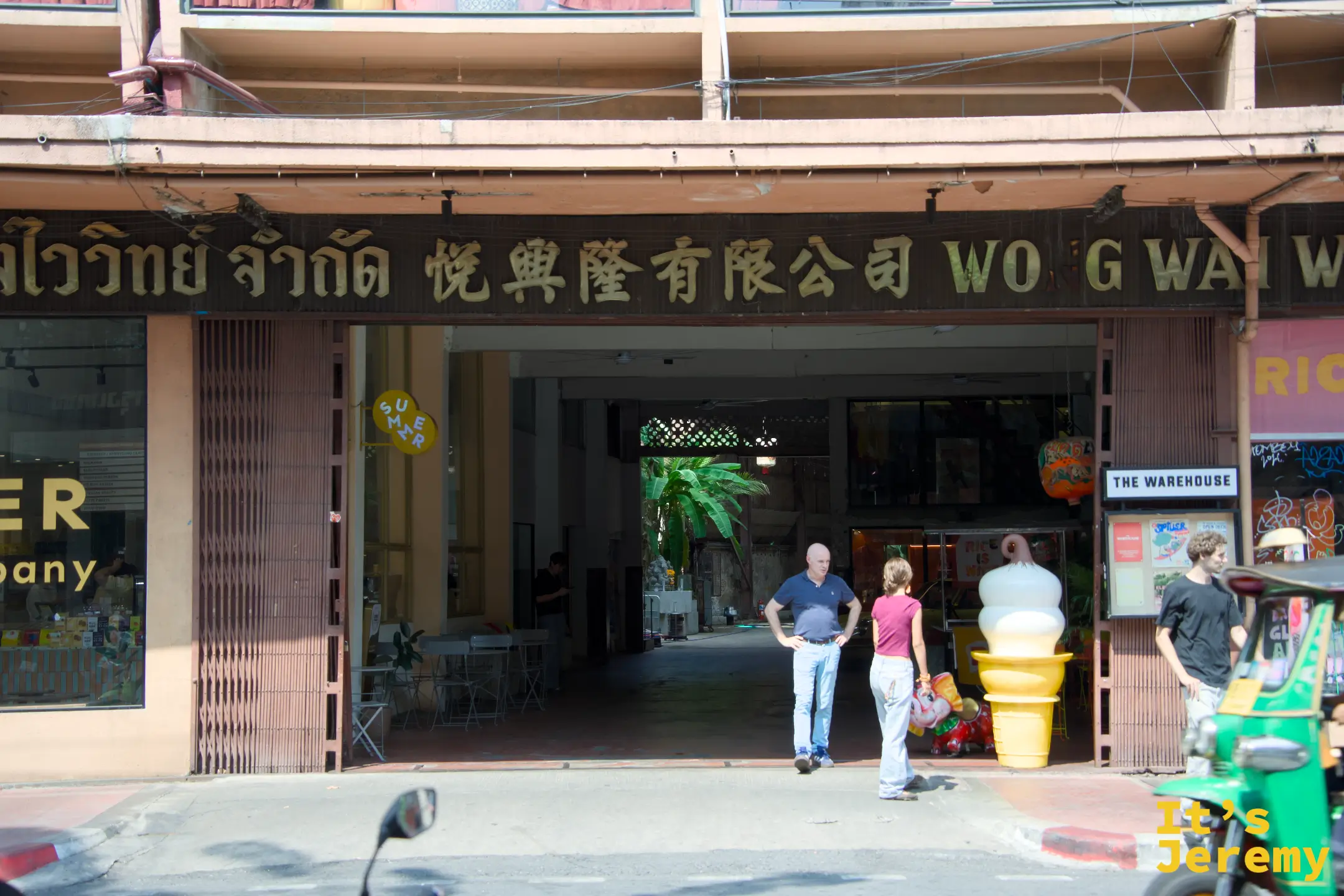 THE WAREHOUSE Bangkok 入口外觀:老倉庫門洞,旁邊掛著「悅興隆有限公司」招牌,THE WAREHOUSE 招牌在斜後方,一台嘟嘟車停在外