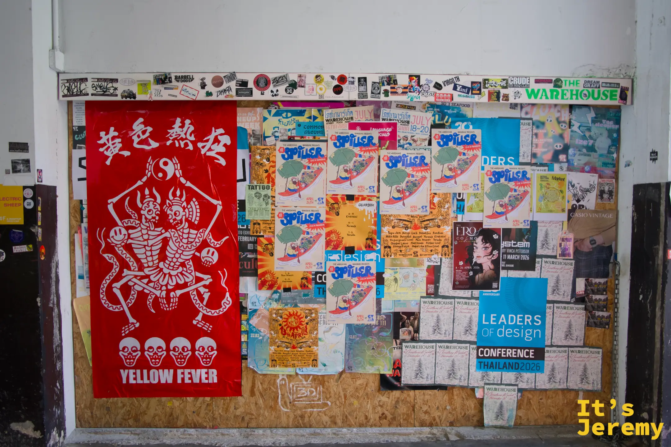 公佈欄上貼著「黃色熱狂 Yellow Fever」展覽旗幟和多張活動海報,上方角落有「THE WAREHOUSE」字樣