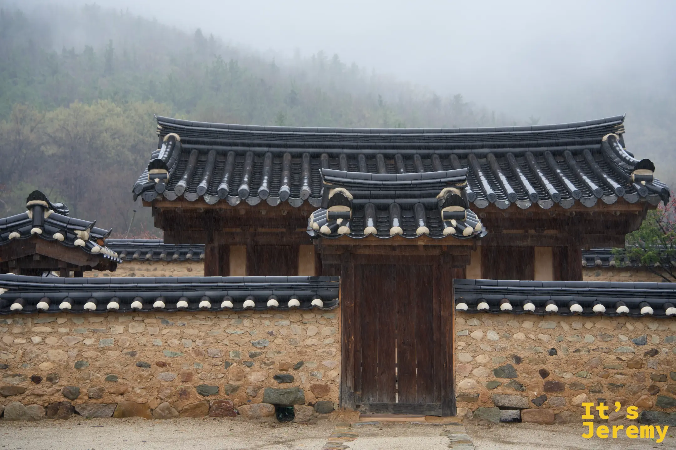 雨中的漆谷韓屋村正門，青瓦大門後是霧氣瀰漫的山，雨絲清晰可見