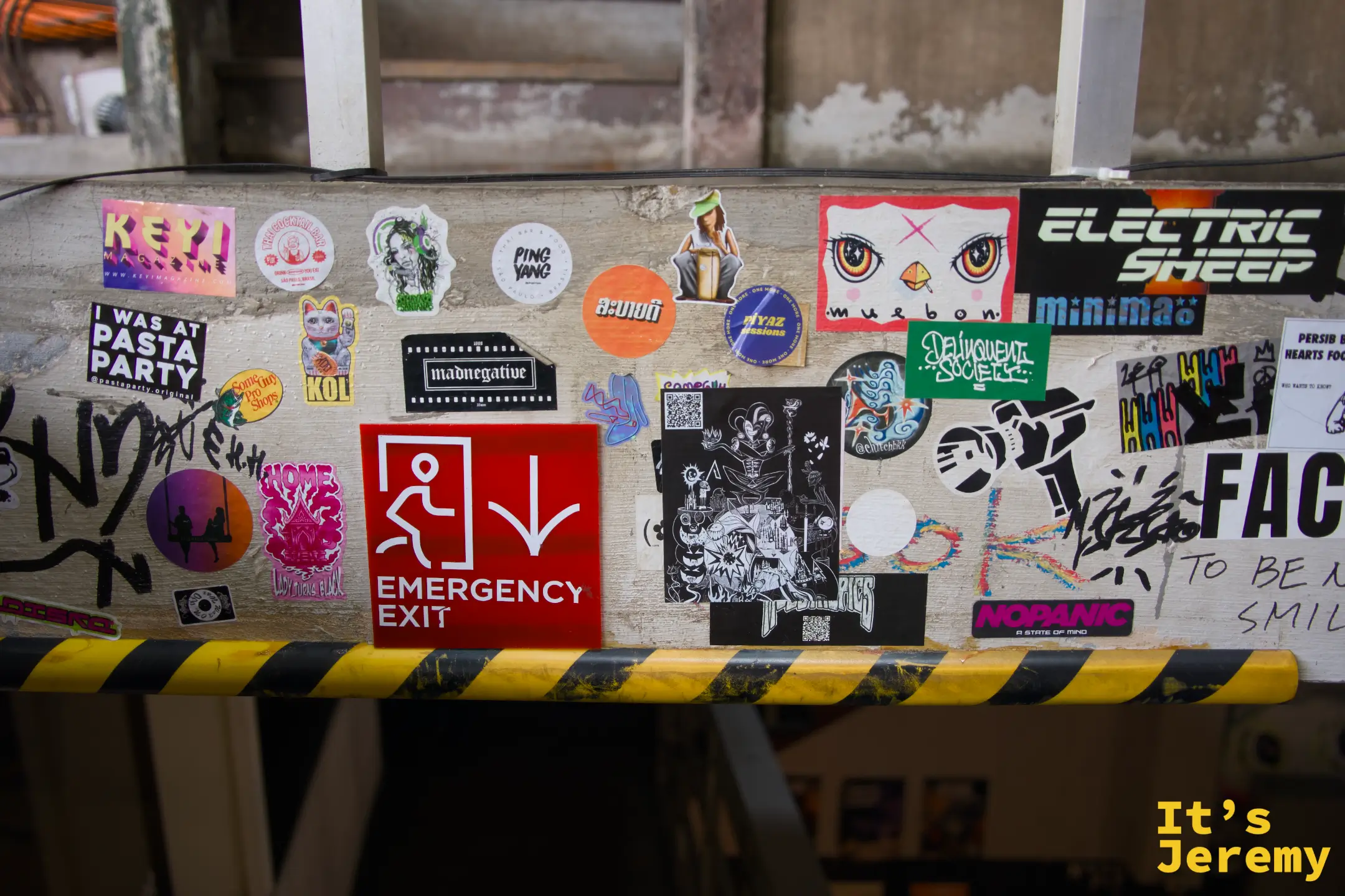 橫梁上密布貼紙:Emergency Exit 紅色標誌夾在各種品牌貼紙之間,包括 Electric Sheep、FACH 字樣