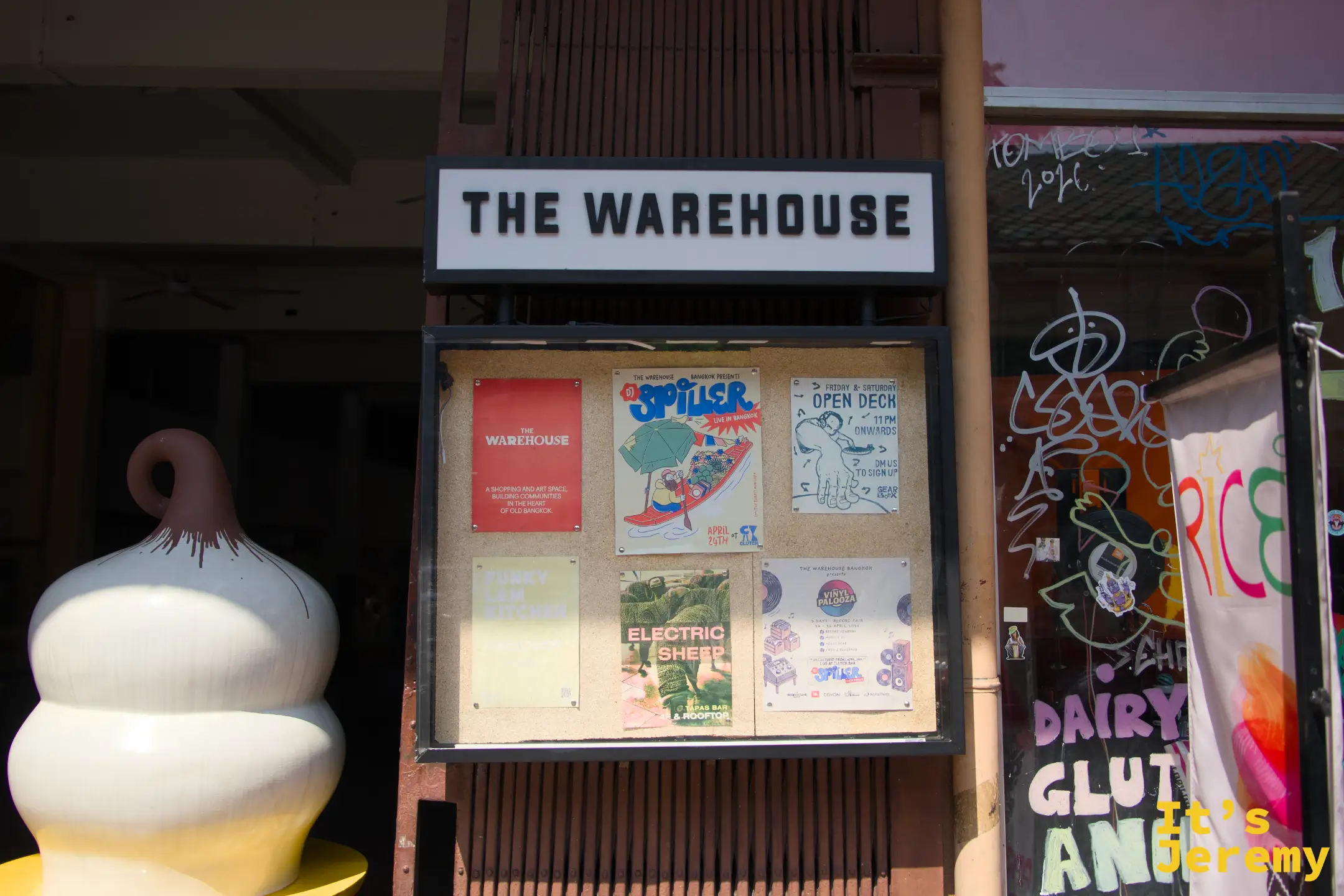 THE WAREHOUSE 正面外觀:「THE WAREHOUSE」白字招牌,下方是活動佈告欄,旁邊有白色陶瓷裝置藝術