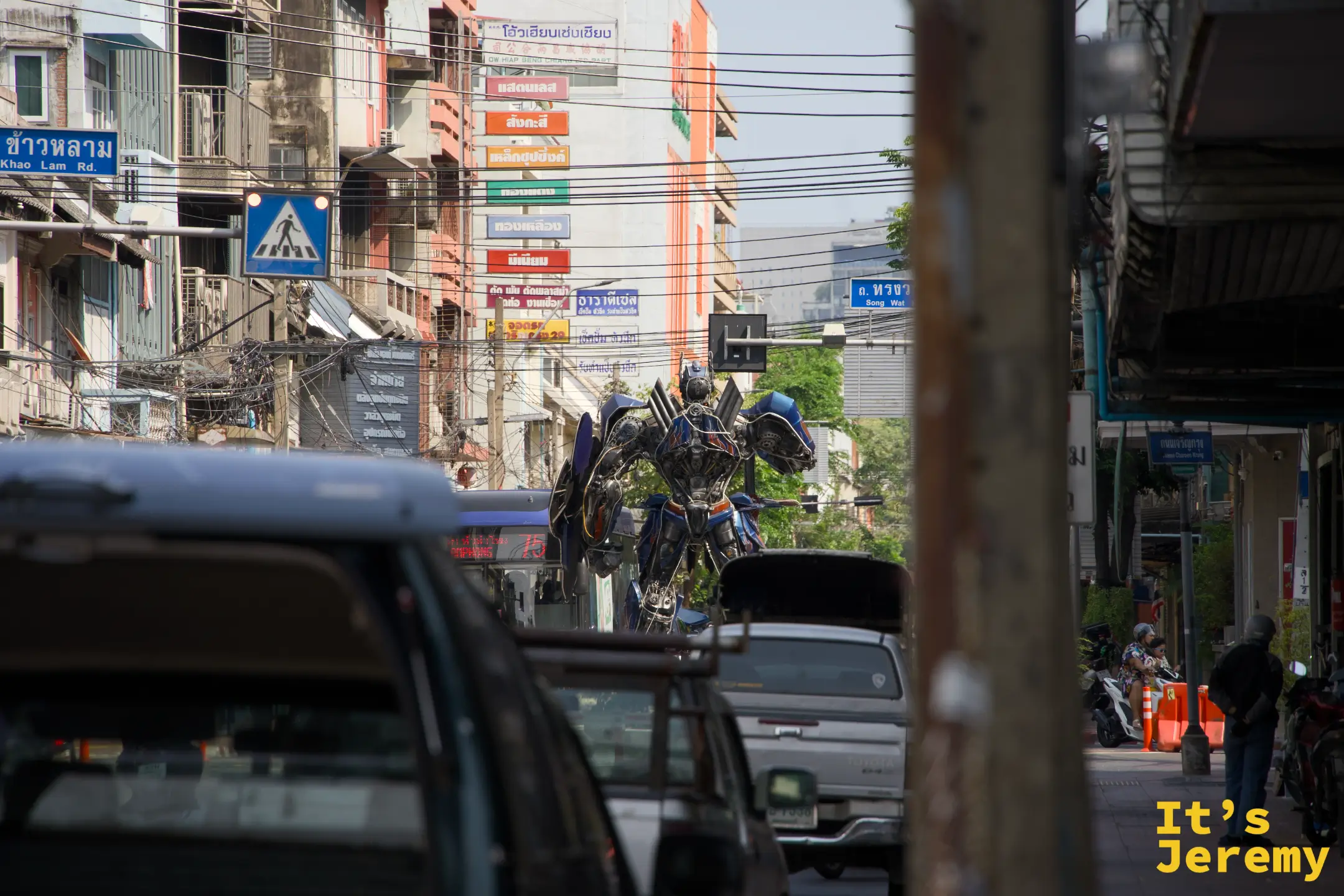 從遠處透過車流看到嵩越路路口,擎天柱雕塑在車輛之間矗立,路牌標示「Khao Lam Rd.」和「Song Wat」