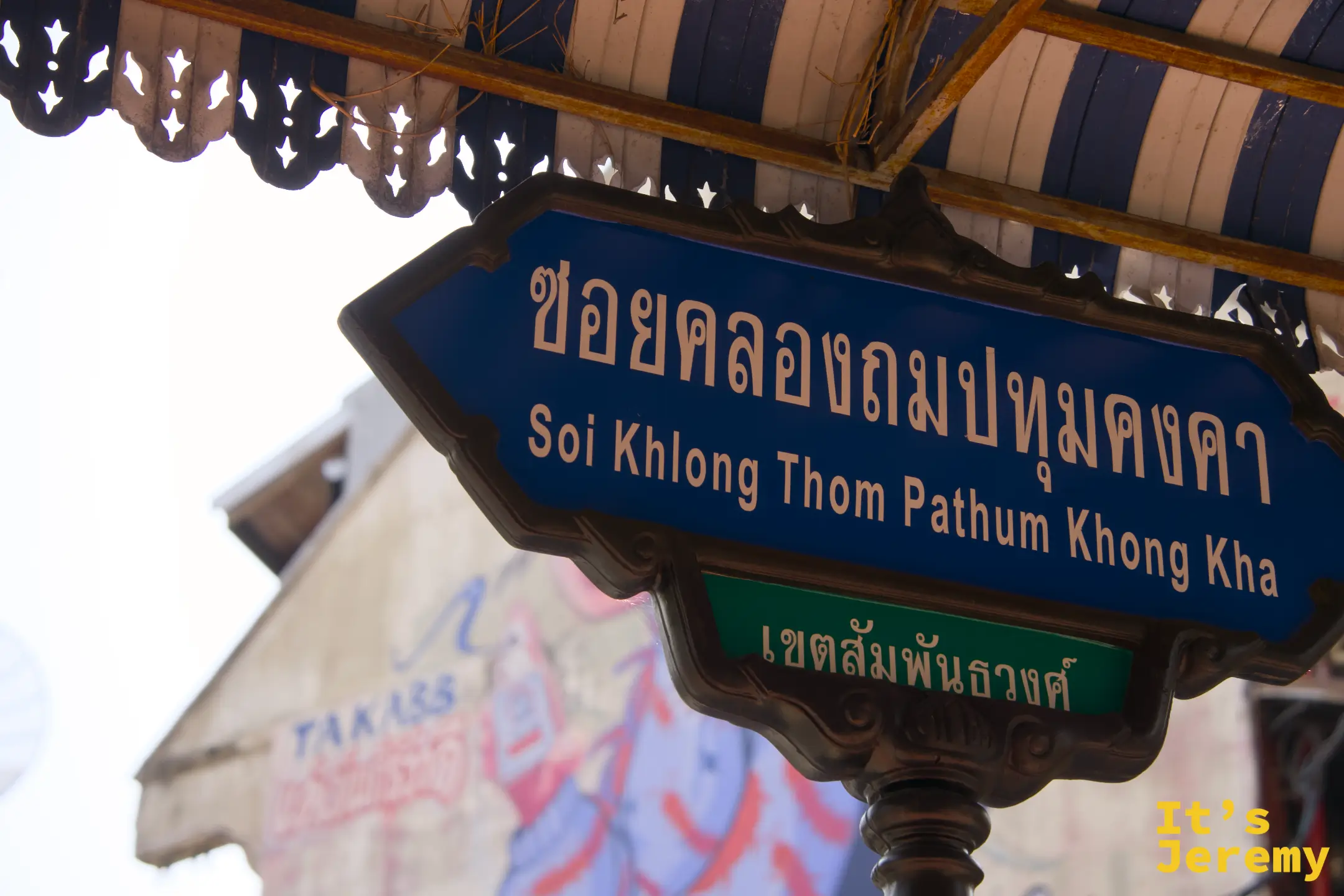 藍色泰英雙語路標「Soi Khlong Thom Pathum Khong Kha」,背後建築牆面是密集的塗鴉字體