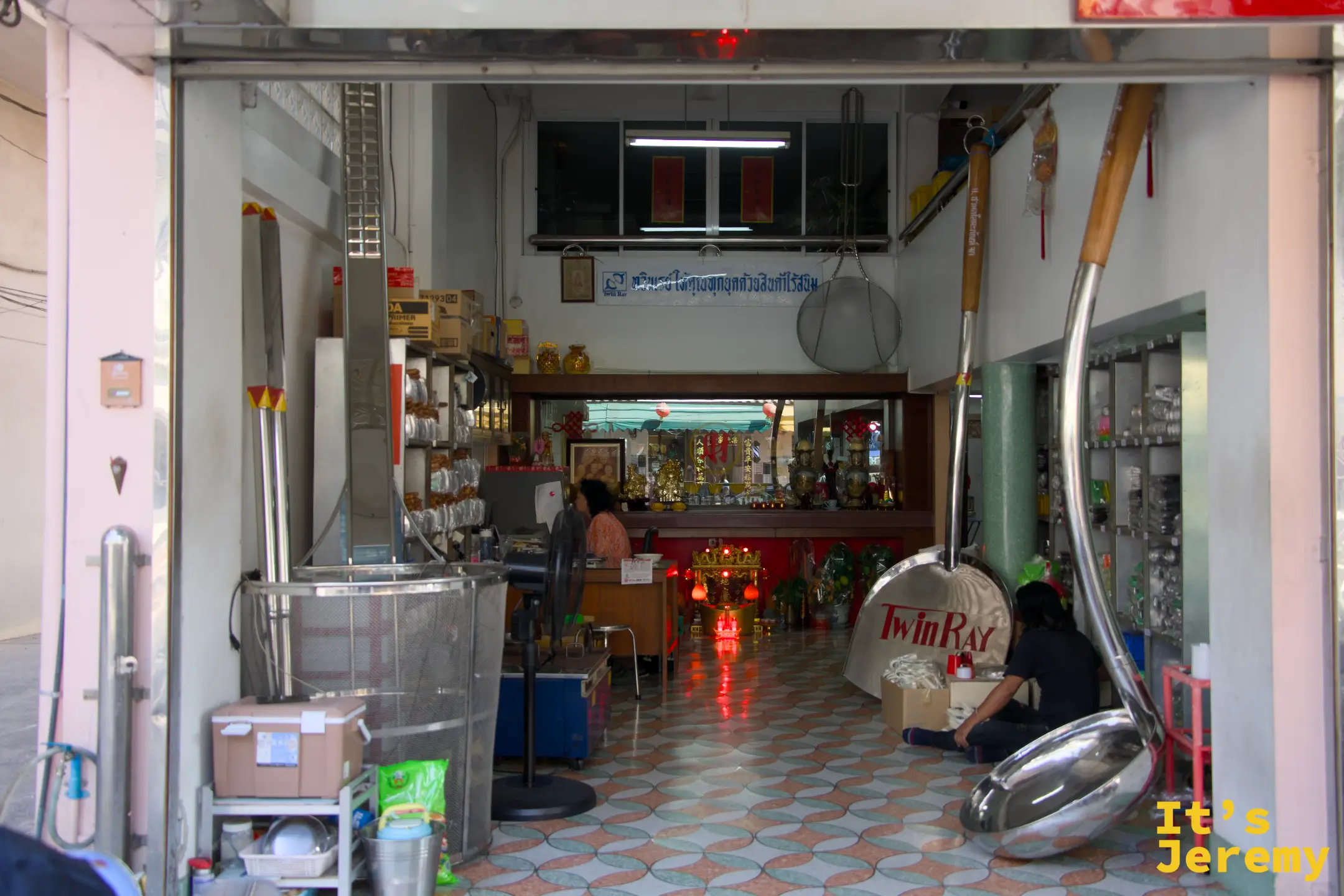 廚具行入口:超大型不鏽鋼炒鏟和舀勺靠在門邊,店內可見神桌、磁磚地板和女店員
