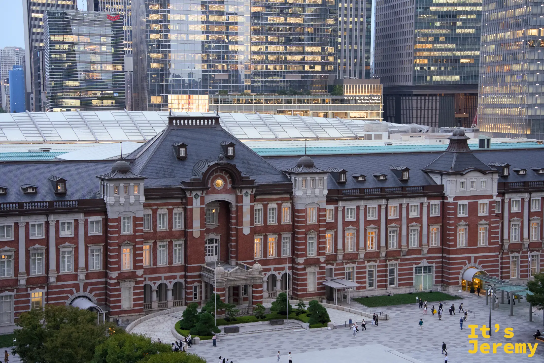 傍晚從高處俯拍丸之內廣場，東京車站站體完整展開，背後摩天大樓群隱入暮色