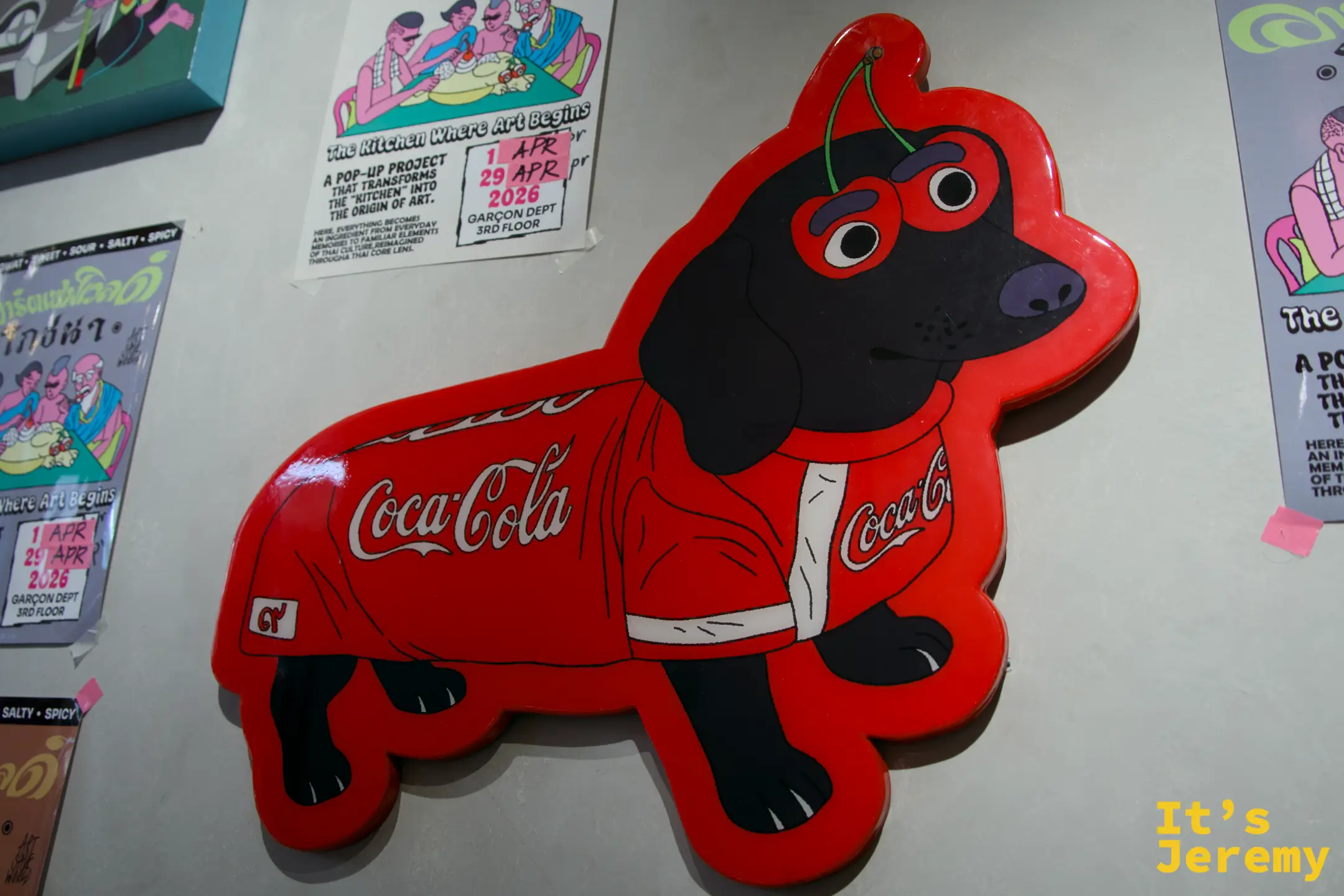 黑色臘腸犬穿著可口可樂球衣的牆面裝置藝術，旁邊貼著「The Kitchen Where Art Begins」展覽資訊
