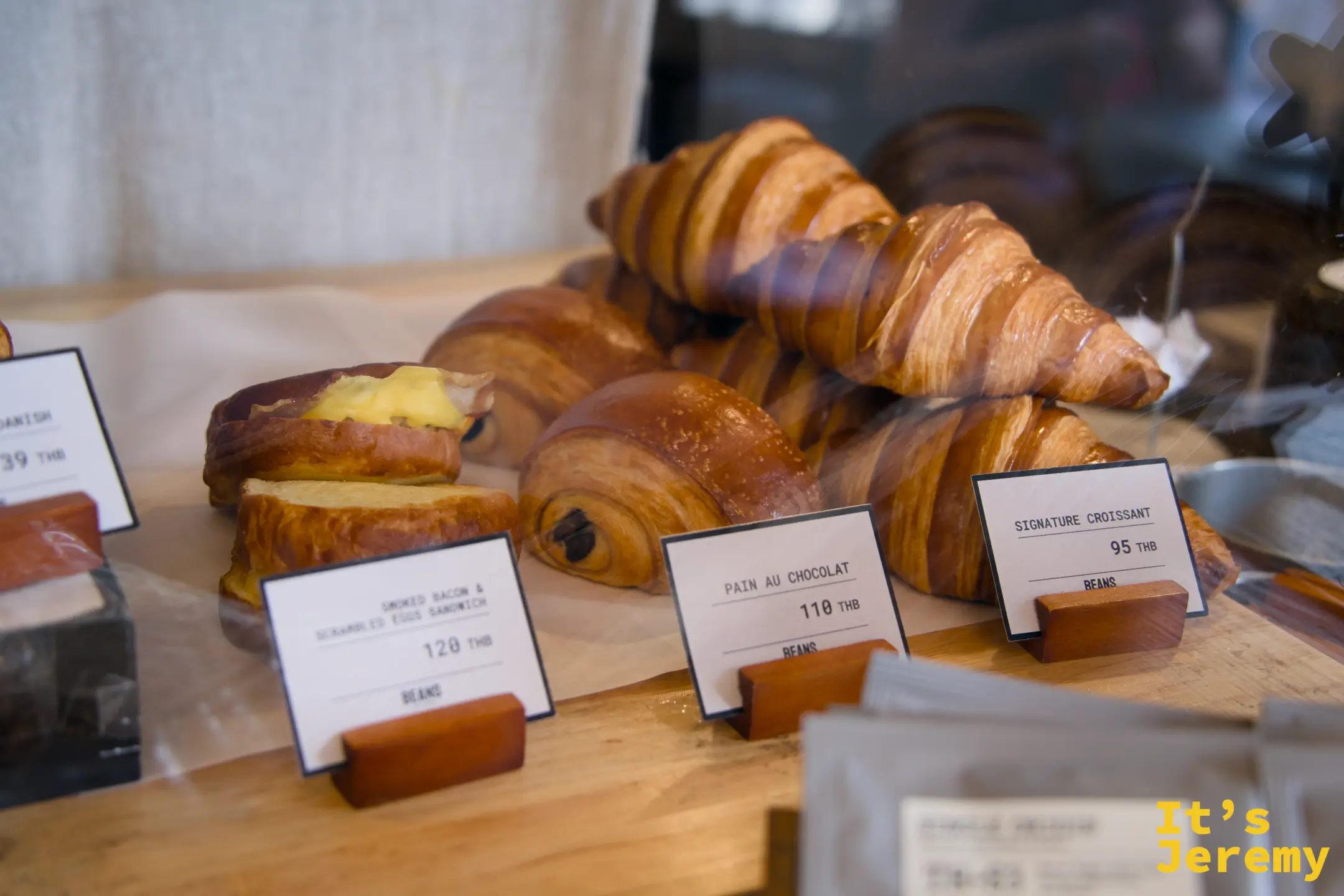 玻璃展示櫃中的甜點:Signature Croissant ฿95、Pain au Chocolat ฿110、Smoked Bacon & Scrambled Eggs Sandwich ฿120、Sausage Danish ฿39