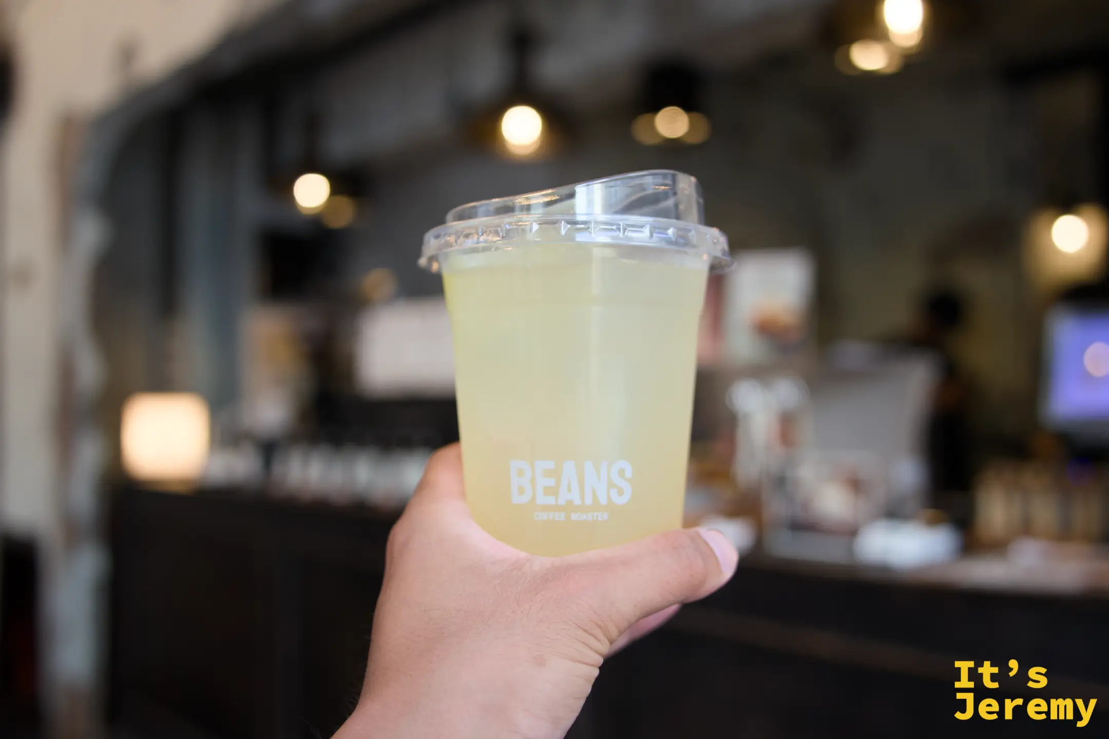 手持 BEANS 品牌透明杯,杯內為淡黃綠色柚子蘇打(Yuzu Soda),店內燈光與裸磚牆作為背景