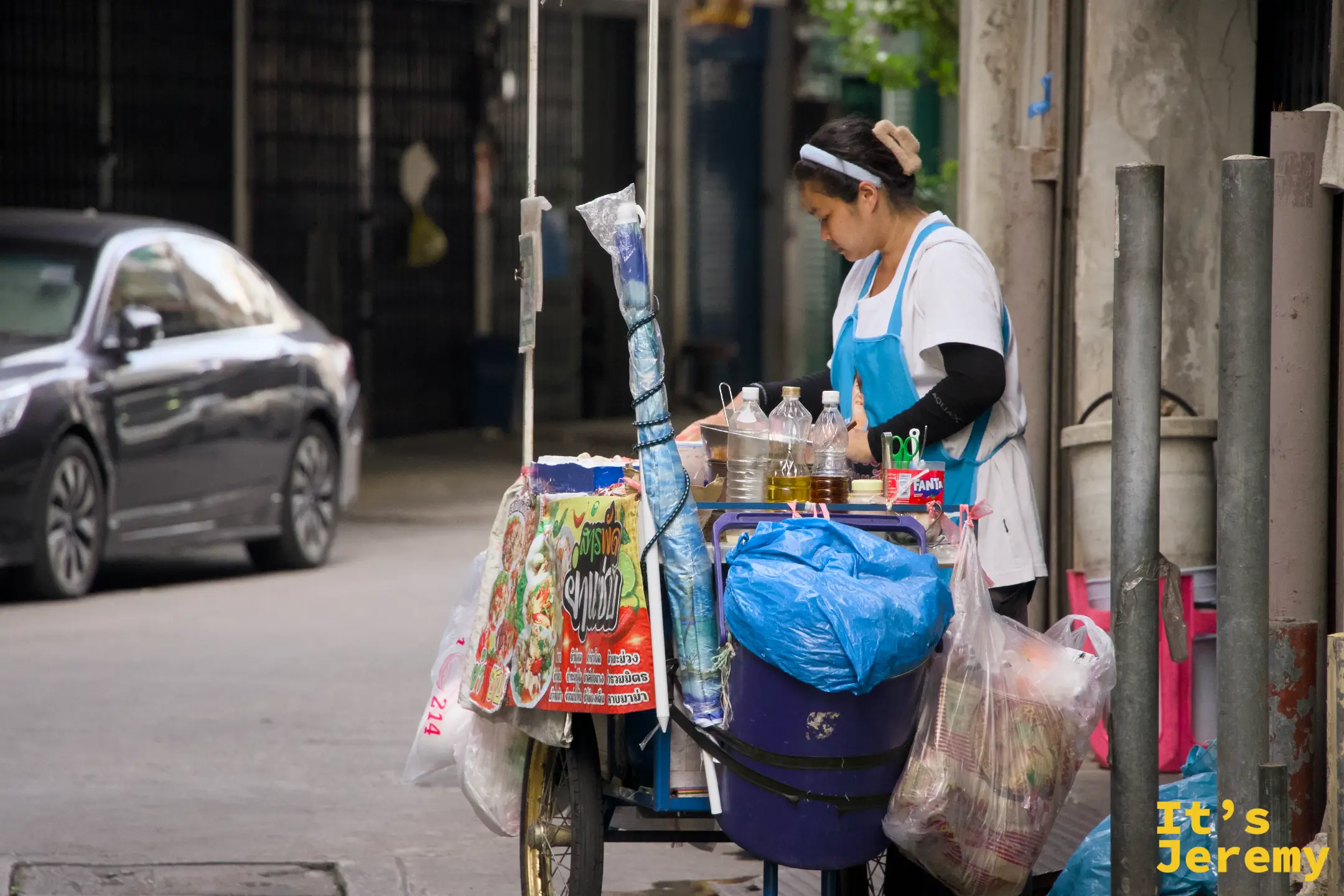 女攤主在推車旁整理食材和袋裝物品,推車上有各式飲料和調味料