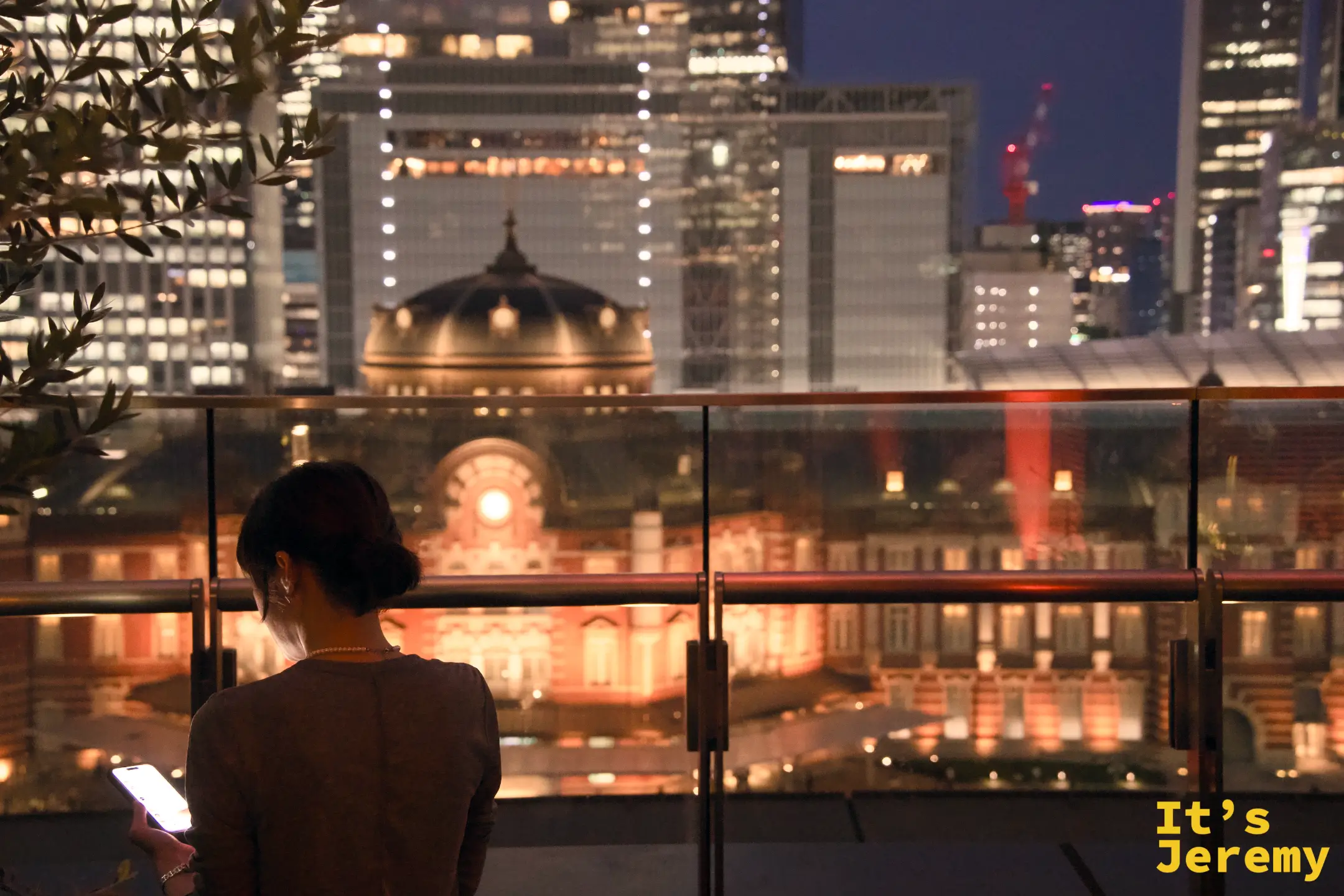 露台女性背影，東京車站橘紅夜景在玻璃護欄上透出倒影