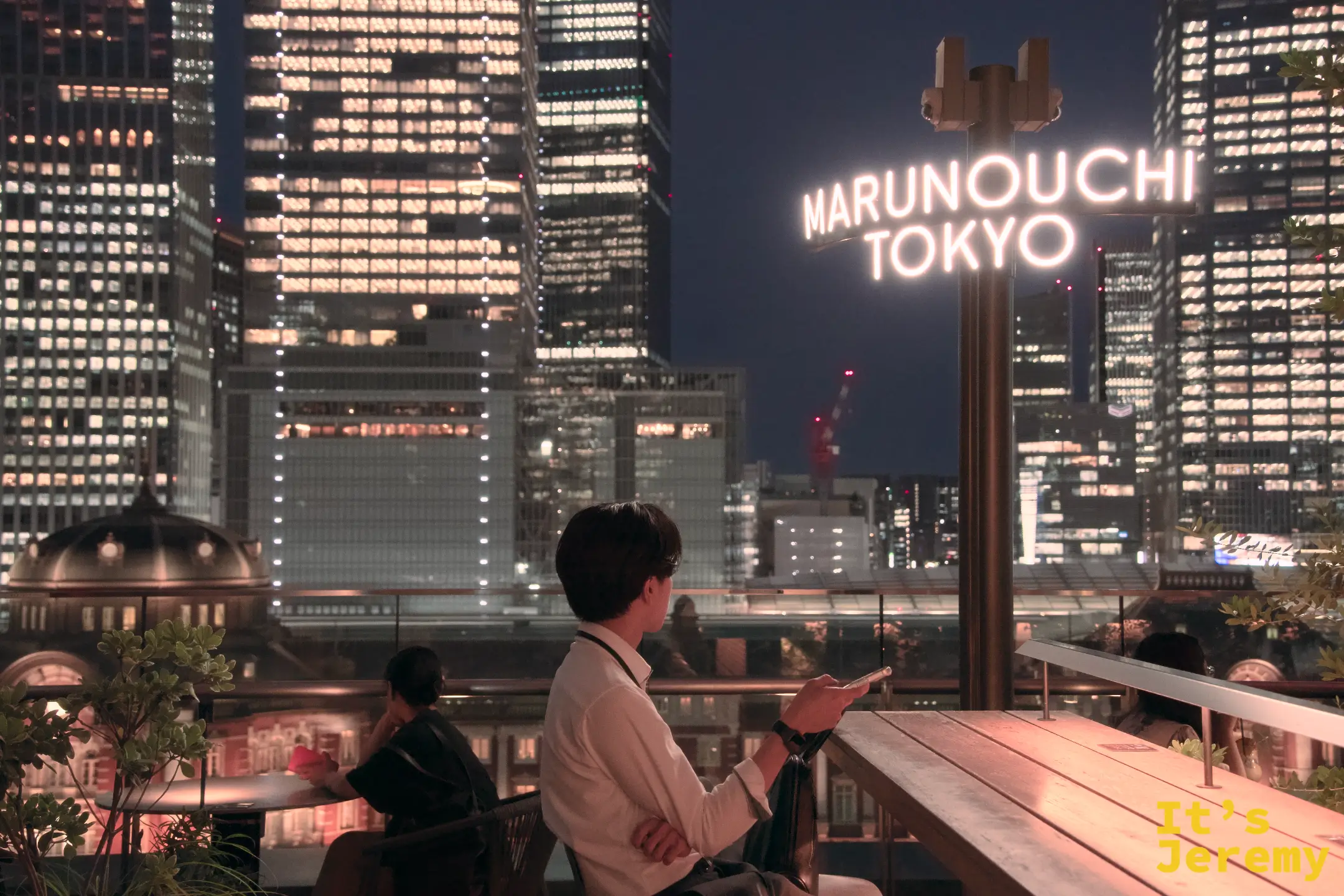 夜晚露台上，男性靠著 MARUNOUCHI TOKYO 霓虹燈牌，背後是整個丸之內的入夜景色