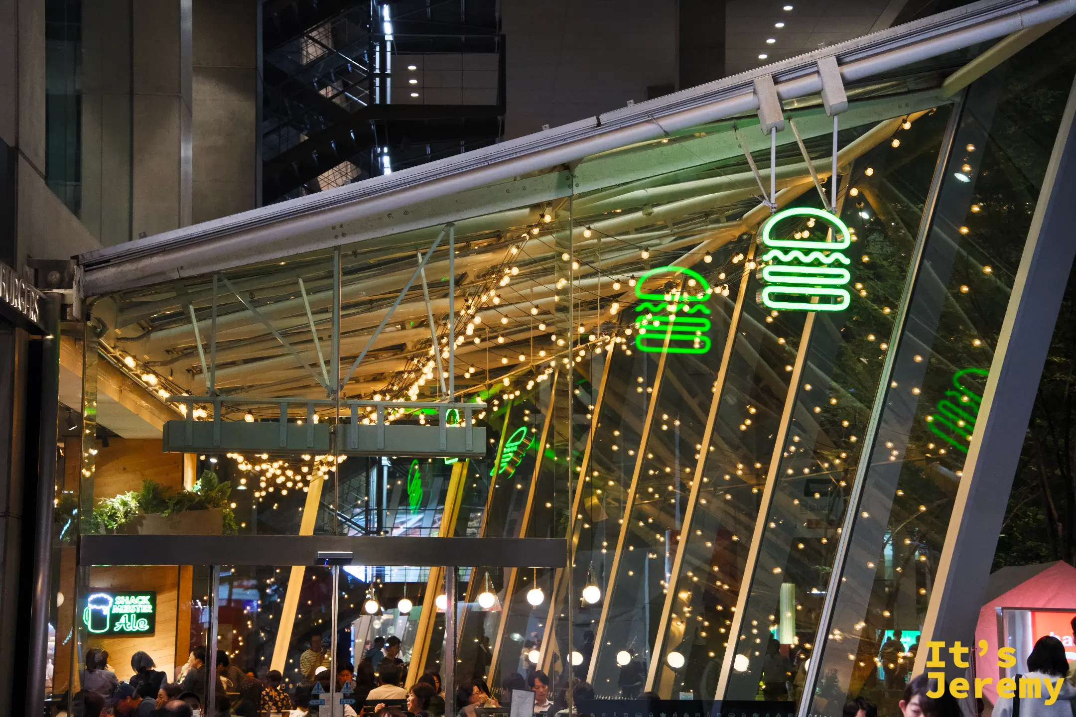 東京國際論壇廣場夜景,Shake Shack 戶外座位區在串燈與霓虹漢堡 logo 的映照下,客人陸續入座