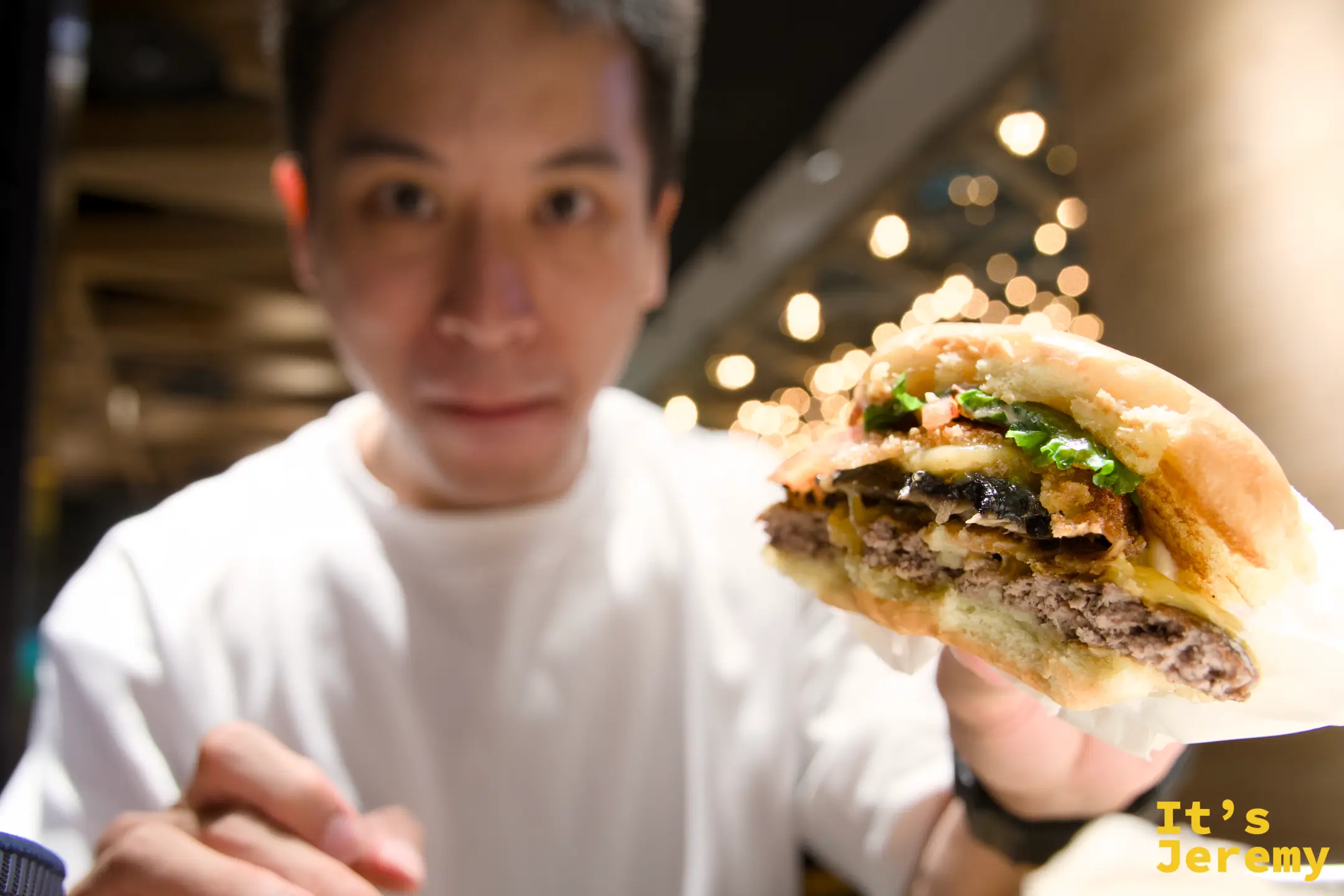 Jeremy 手持咬了一口的 ShackStack 漢堡,截面清楚顯示牛肉、起司、炸波特貝羅蘑菇的三層結構,背景是餐廳暖色燈光 bokeh