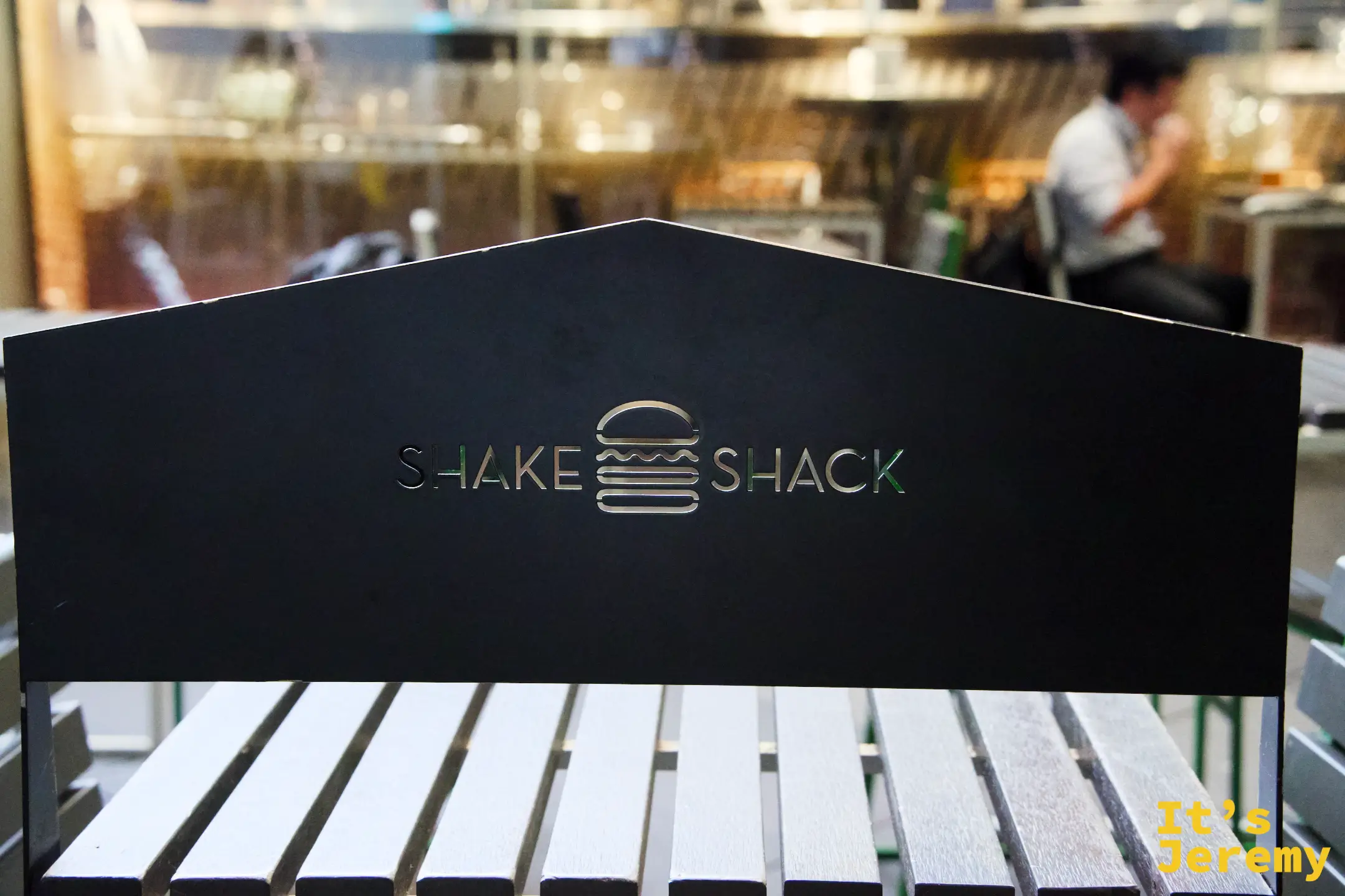 戶外廣場木質長椅椅背上的金色 Shake Shack logo,背景是餐廳室內的暖色燈光