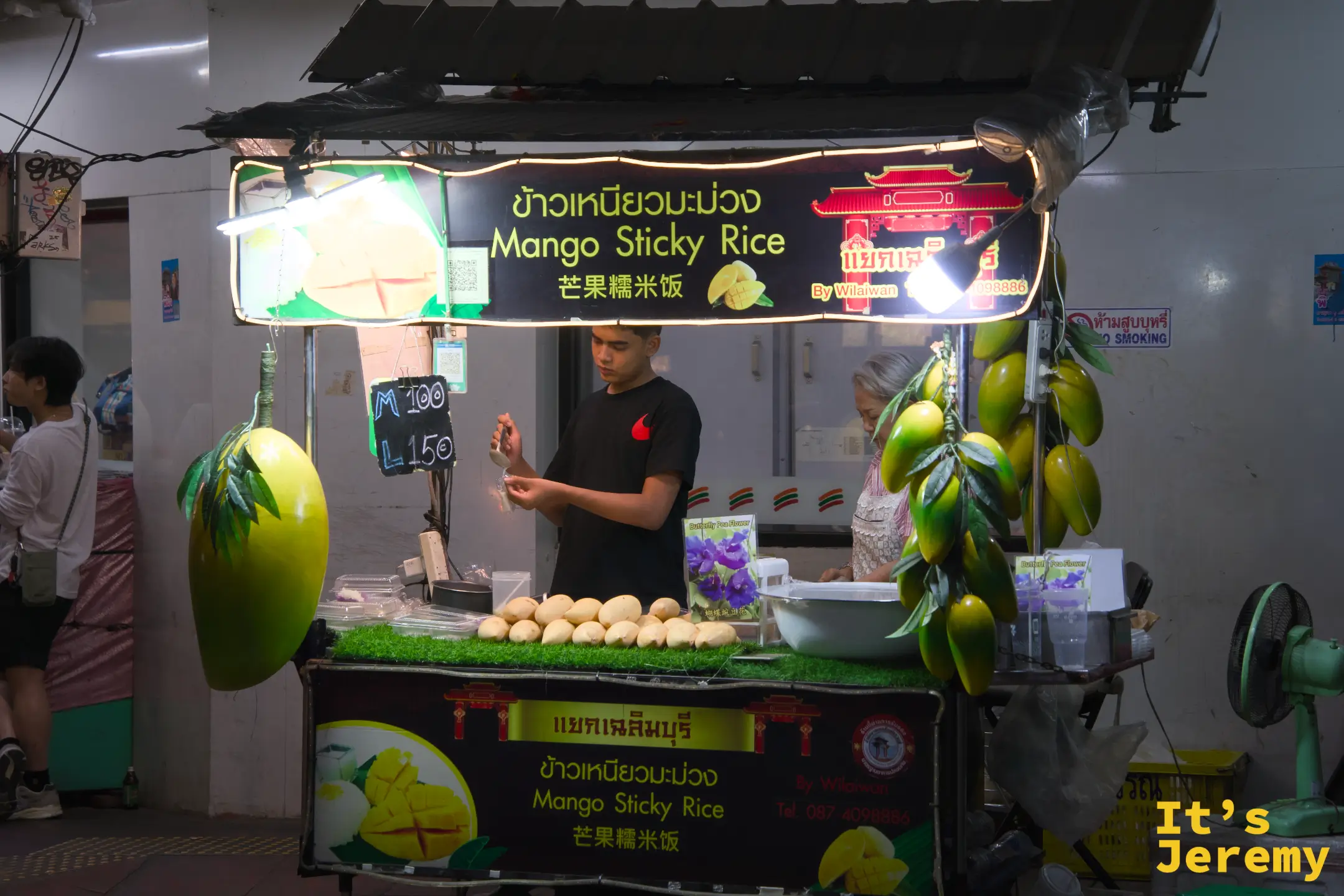 芒果糯米飯夜間小攤:鮮綠色假芒果掛飾、白熾燈照明,一名男生在整理糯米