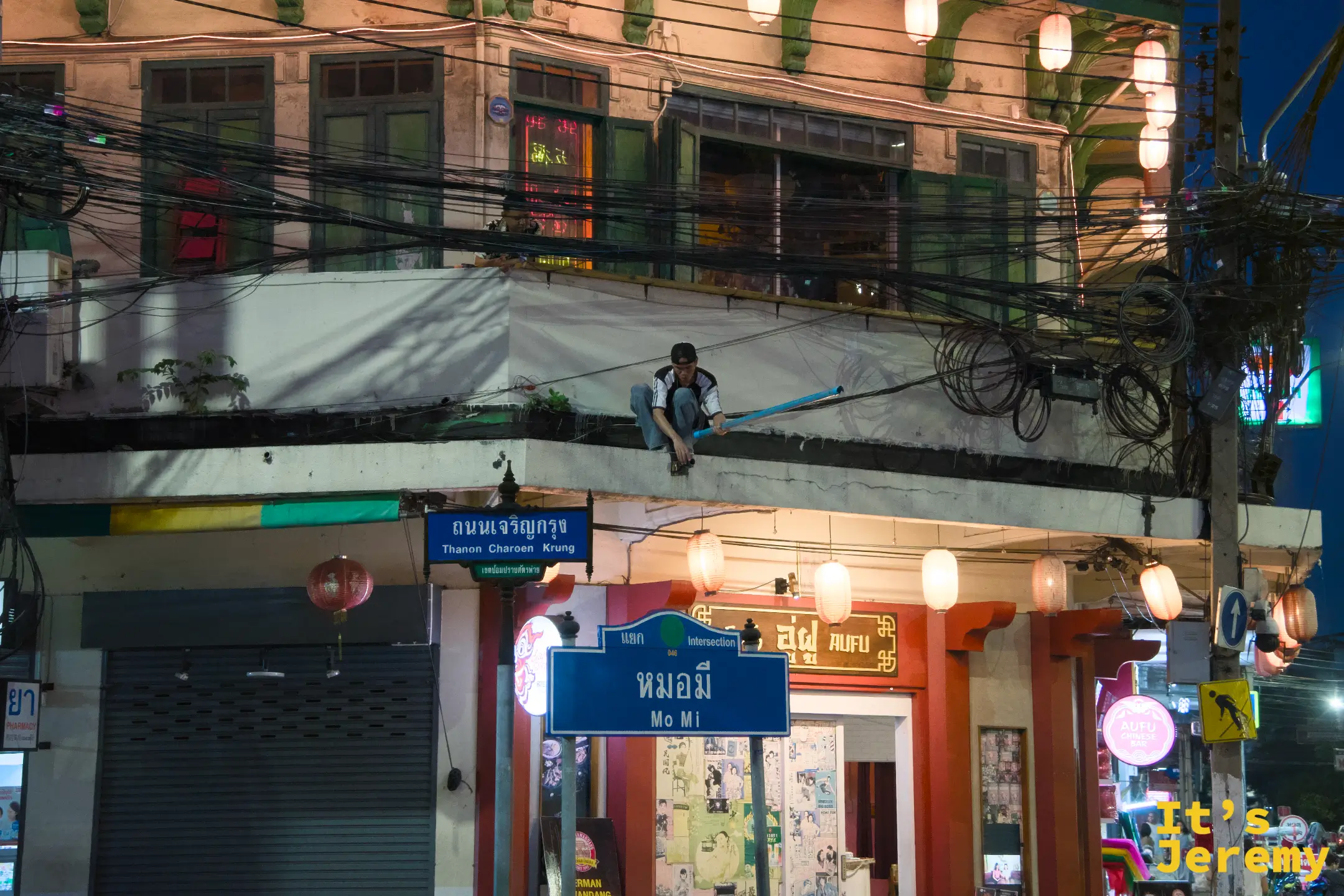 夜間路口:「Thanon Charoen Krung」路牌下方是「Mo Mi」小巷指標,一名男子趴在二樓欄杆外,附近食堂有紙燈籠