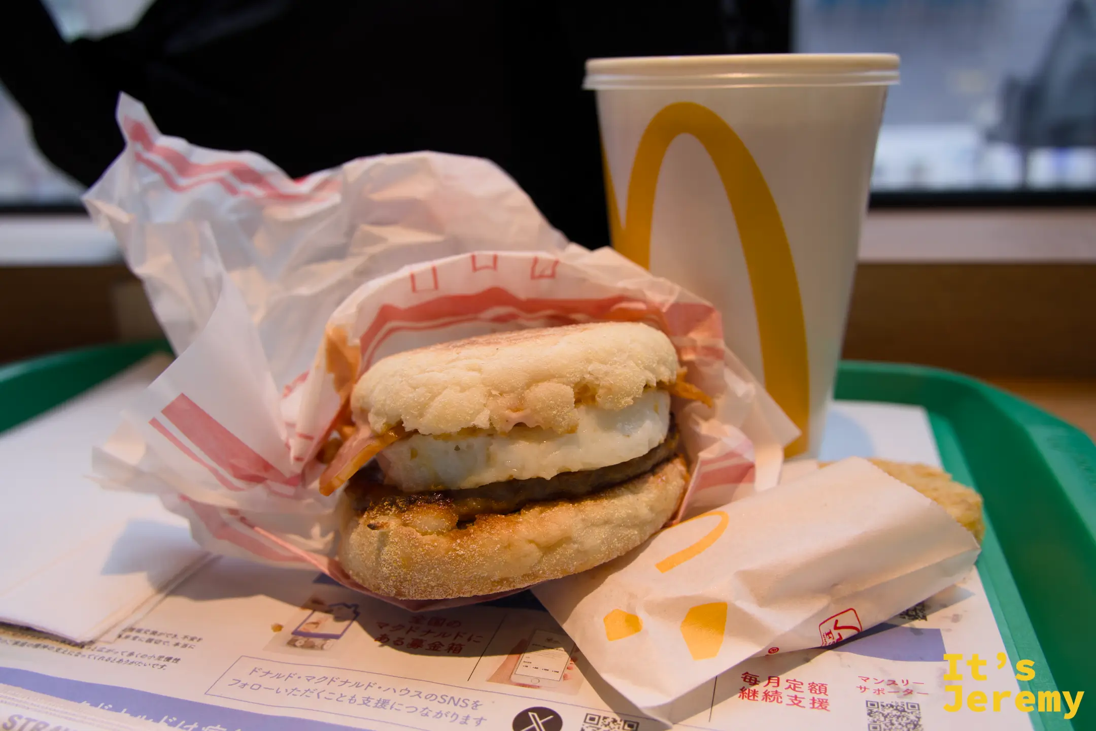 麥當勞早餐:英式鬆餅堡半開包裝放在托盤上,旁邊是紙杯飲料,底部可見日本麥當勞的宣傳單