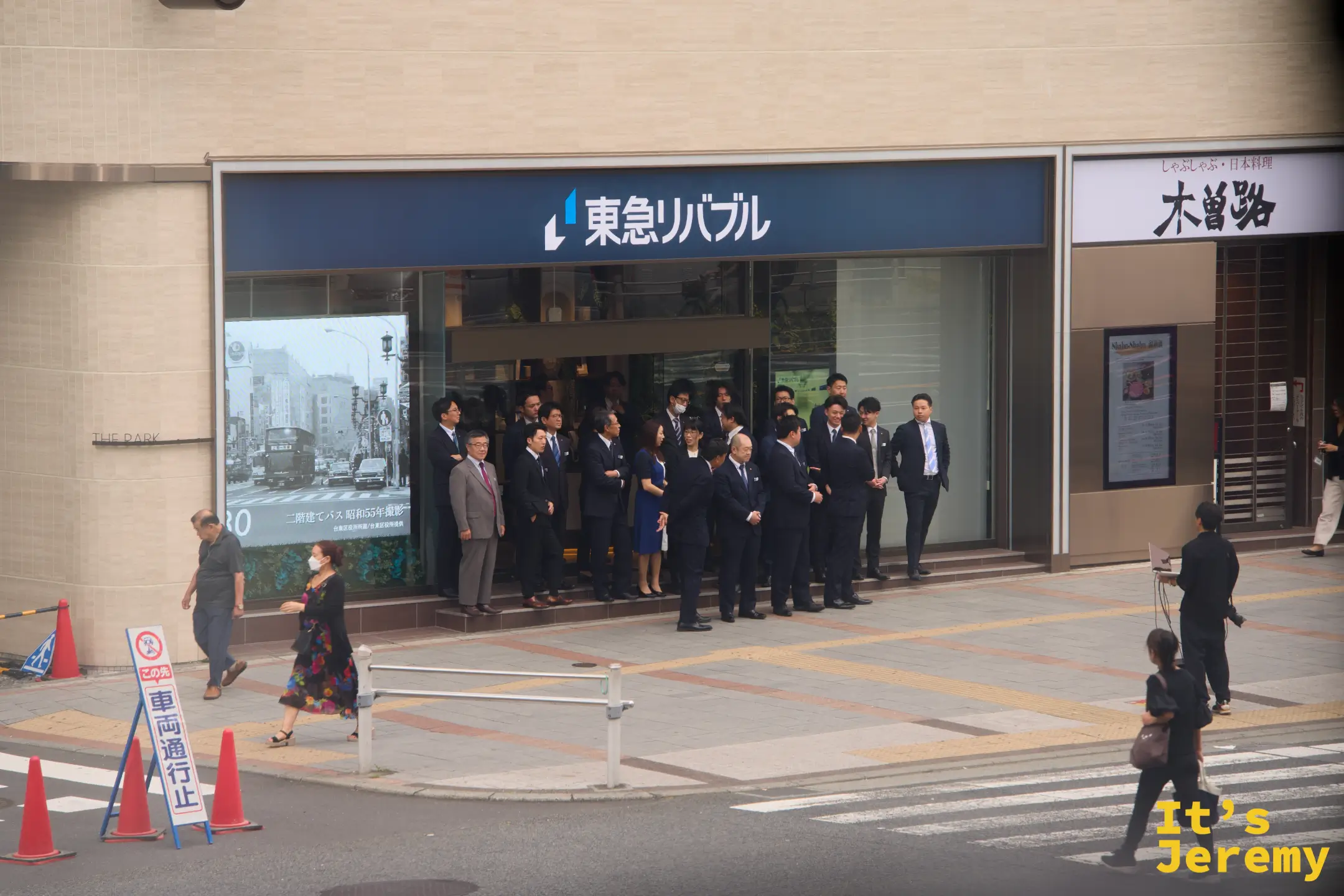 上野附近街道,一群穿西裝的商務人士在東急リバブル房仲店前排隊集合,前方有人捧著電腦充當攝影師,準備拍團體照