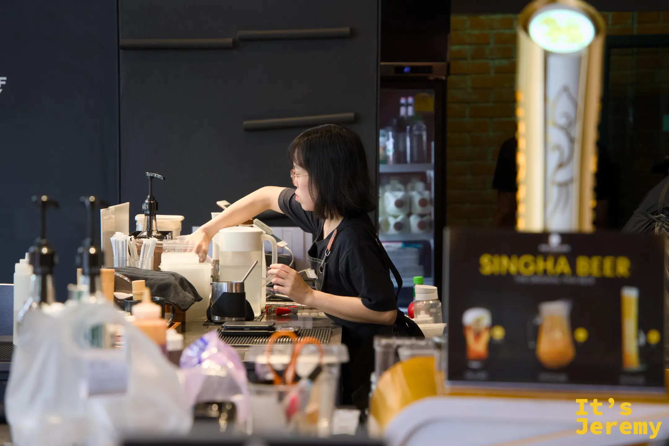 一樓女性咖啡師在吧台工作，側面鏡頭，背後有 Singha Beer 廣告立牌