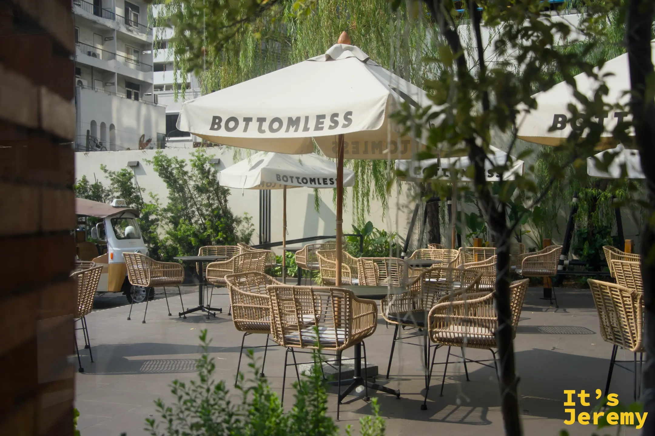 BOTTOMLESS 一樓戶外庭院，白色品牌傘、藤椅與柳樹，有柔和綠意