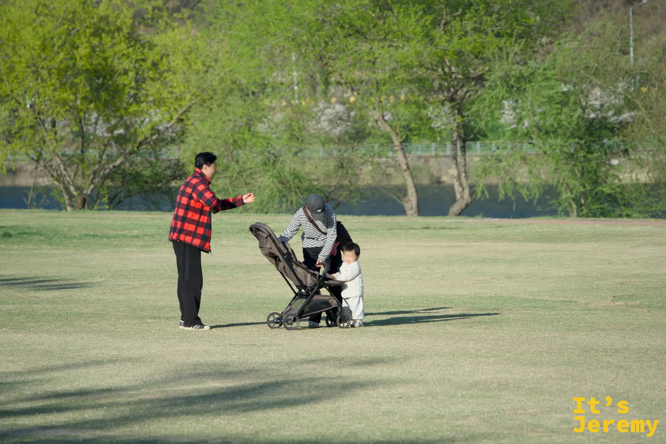 傍晚暖光打在草地上，年輕家庭帶著嬰兒車在江邊慢步，對岸是映著夕照的丘陵
