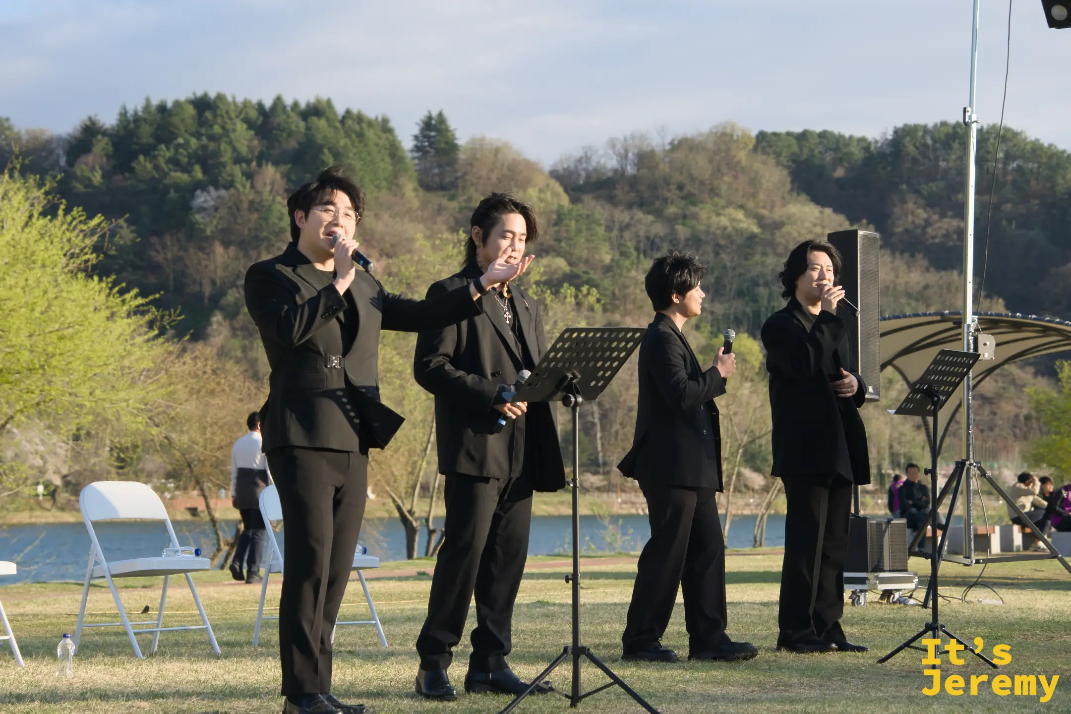四名男聲組合穿黑西裝在江邊草地演出，背景是寬闊的新安川江面與對岸山丘