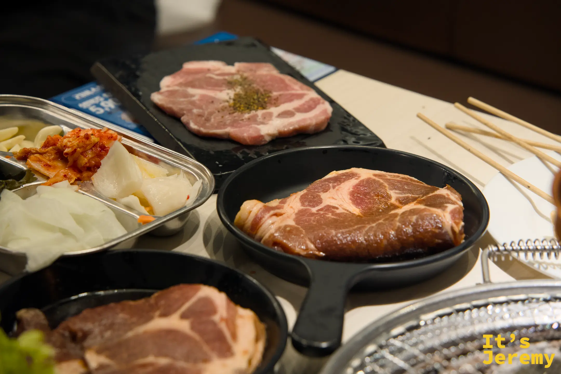 鑄鐵小盤盛著삼겹살與목살，旁邊是整套醃製小菜：大蒜、芝麻葉、泡菜、醃白蘿蔔