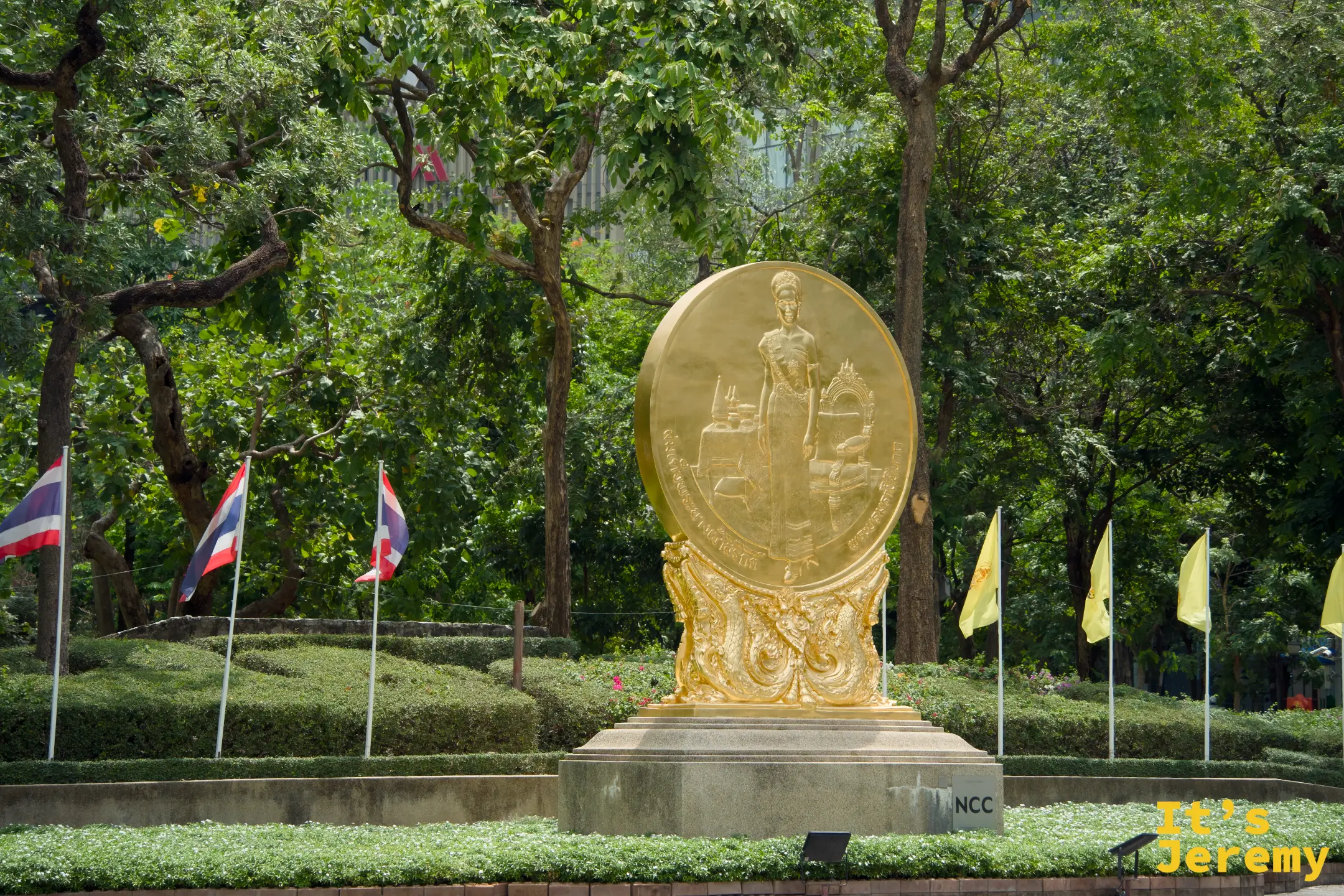 班哲希利公園入口金色圓盤浮雕紀念雕像,周圍有泰國國旗和黃旗,背景為茂密樹木