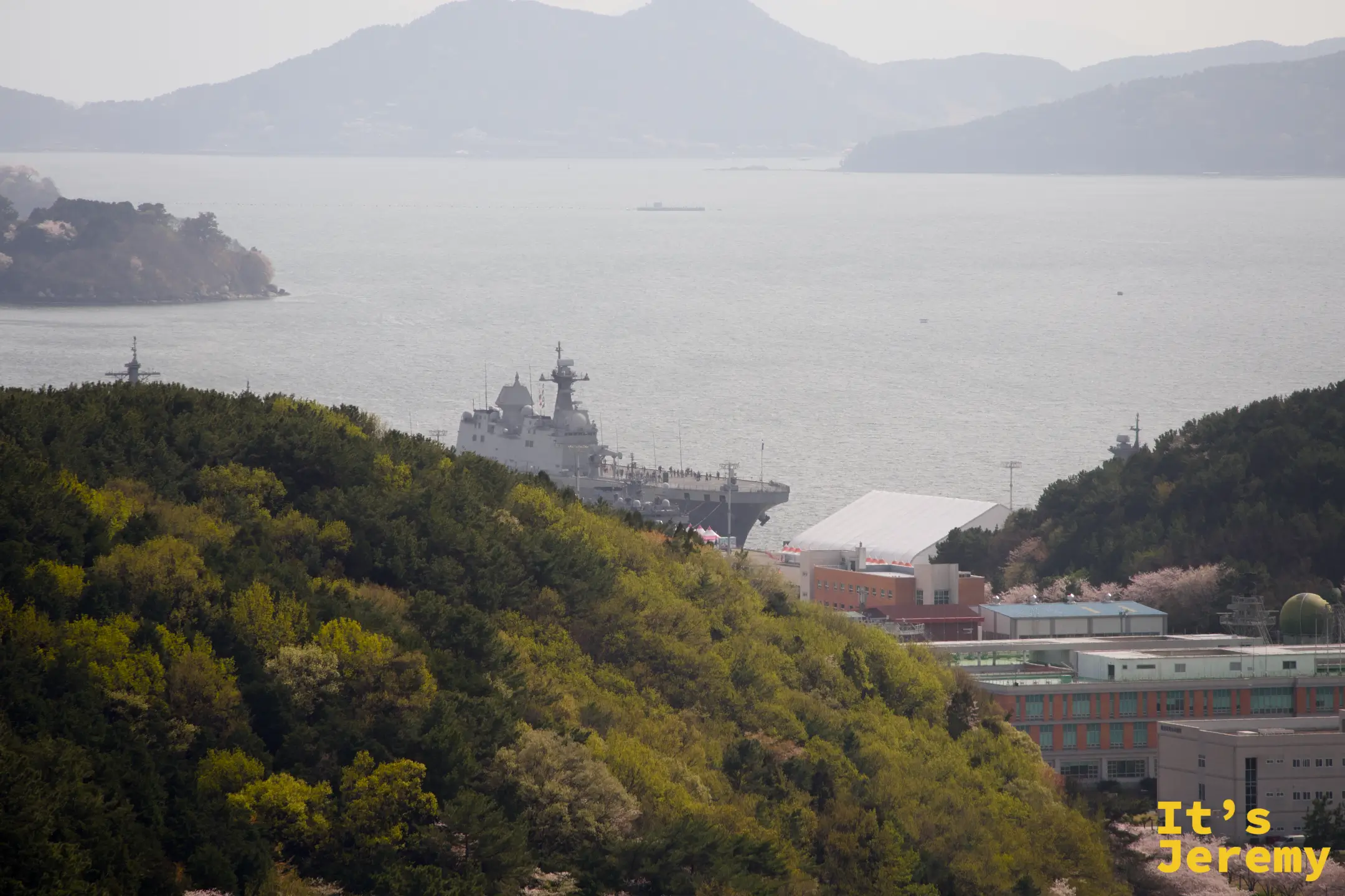港灣後方山丘間停泊的韓國海軍艦艇,與綠色山坡和春日粉白花樹形成強烈對比