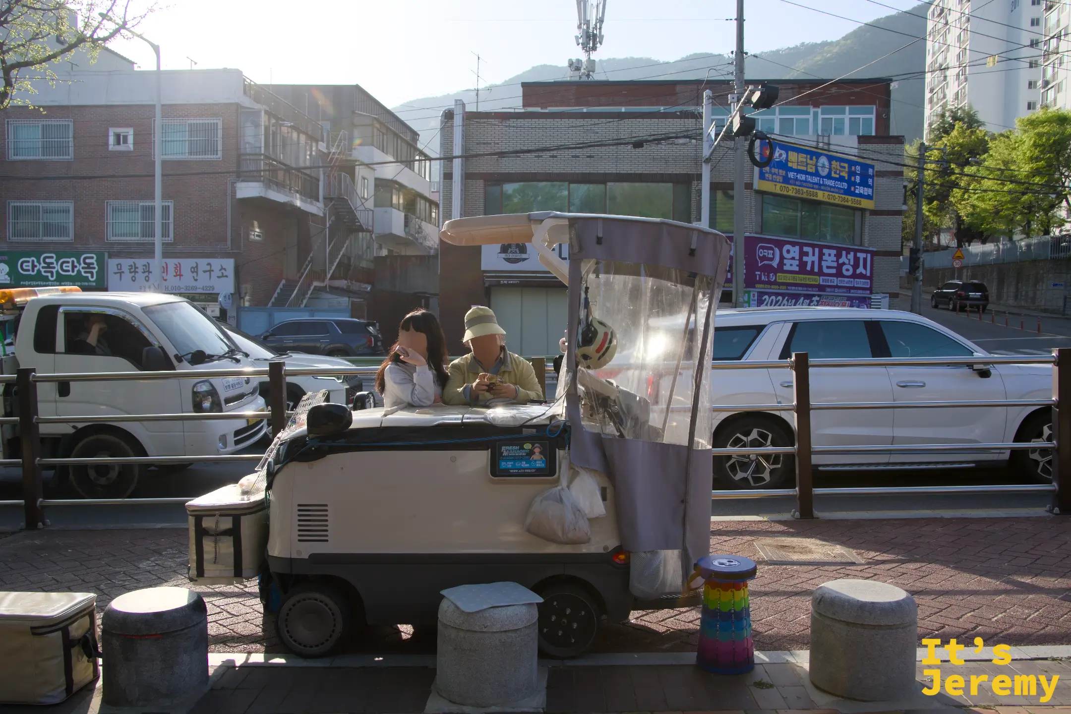 公園外圍街道旁，一輛改裝餐車停在路邊，攤主和女孩並肩在車內，春日斜陽把整個畫面染成金黃