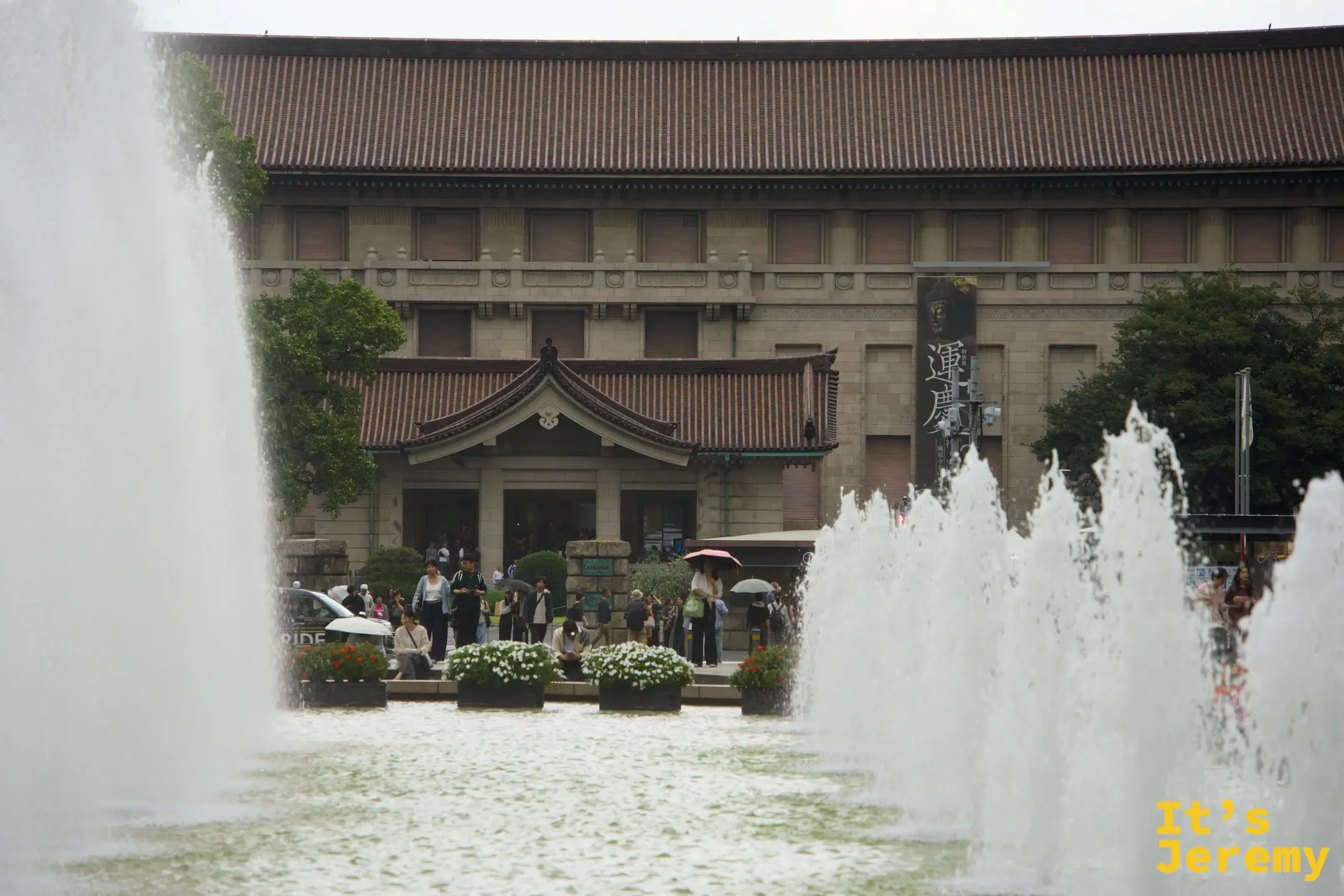 透過噴水池的水霧,東京國立博物館正面建築的日式屋頂清晰可見,廣場前有撐傘的遊客散步其中