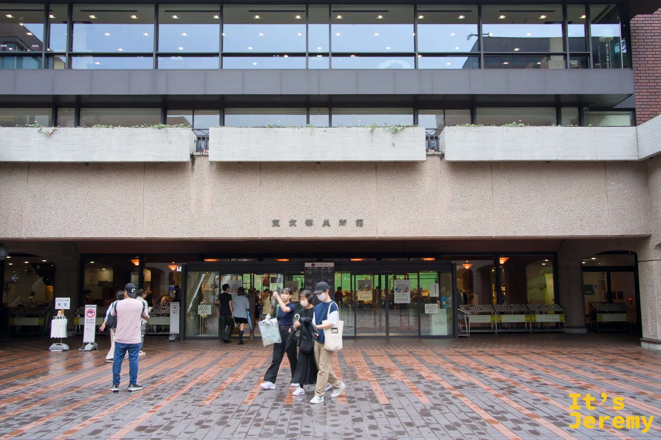 東京都美術館正面入口廣場,磚石地面,「東京都美術館」字樣清晰可見,幾位年輕人從玻璃門魚貫走出